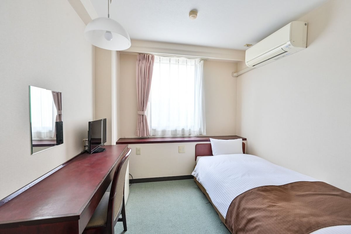 A hotel room with a desk, a bed, a TV, a window with curtains, and an air conditioning unit.