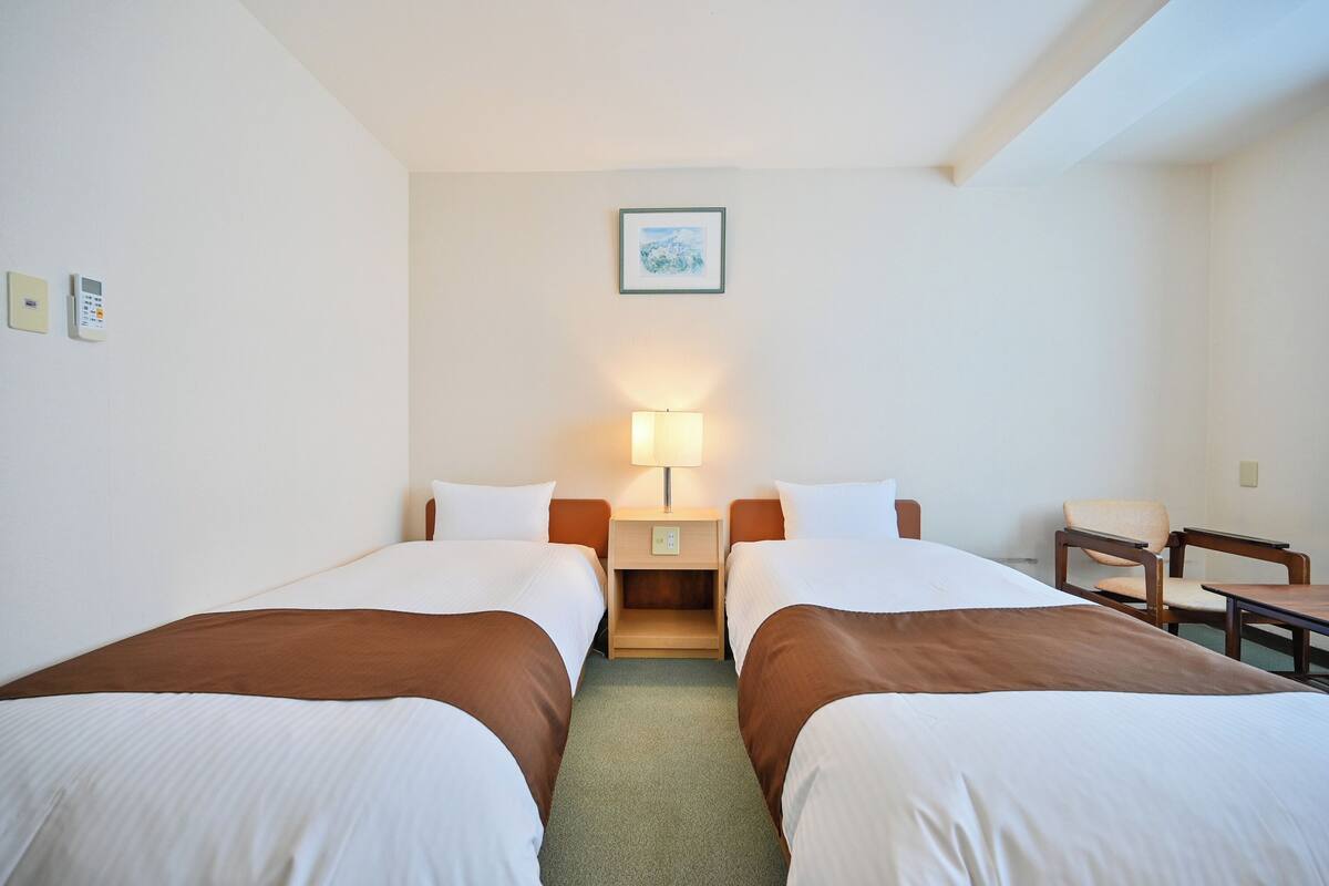 A hotel room with two beds, a nightstand with a lamp, a chair, and a small table.