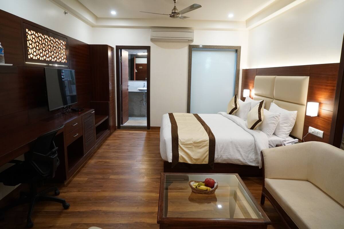 A hotel room with a bed, a desk, a television, a glass coffee table with fruit, and a ceiling fan.