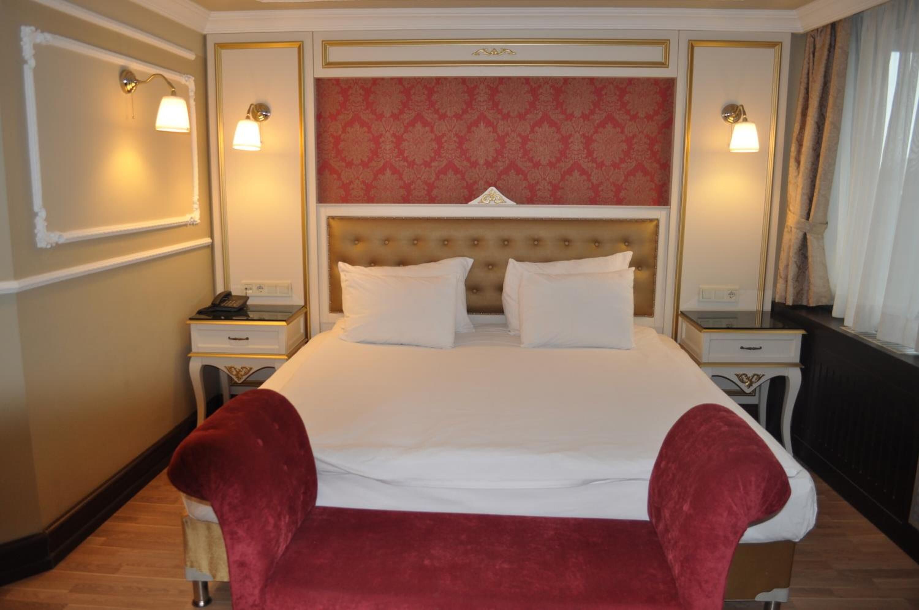 A hotel room with a large bed, two bedside tables, a red upholstered bench, and a patterned wallpaper headboard.