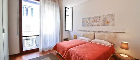 A bedroom with two beds, a large window with curtains, a bedside table with a lamp, and artwork on the wall.