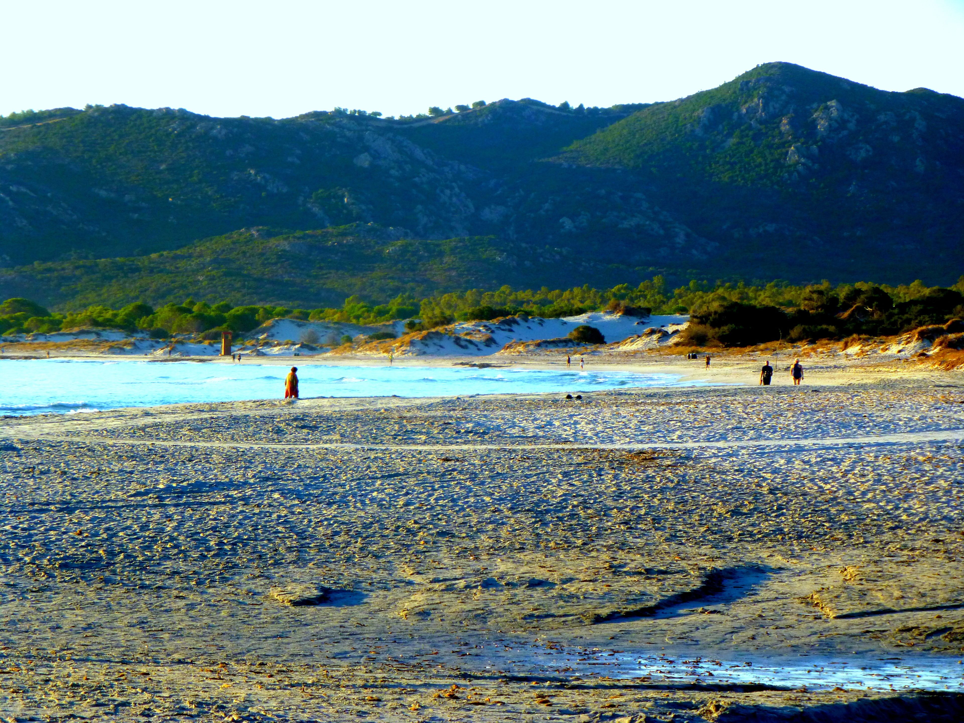 Una playa con piedras, algunas personas y colinas al fondo.