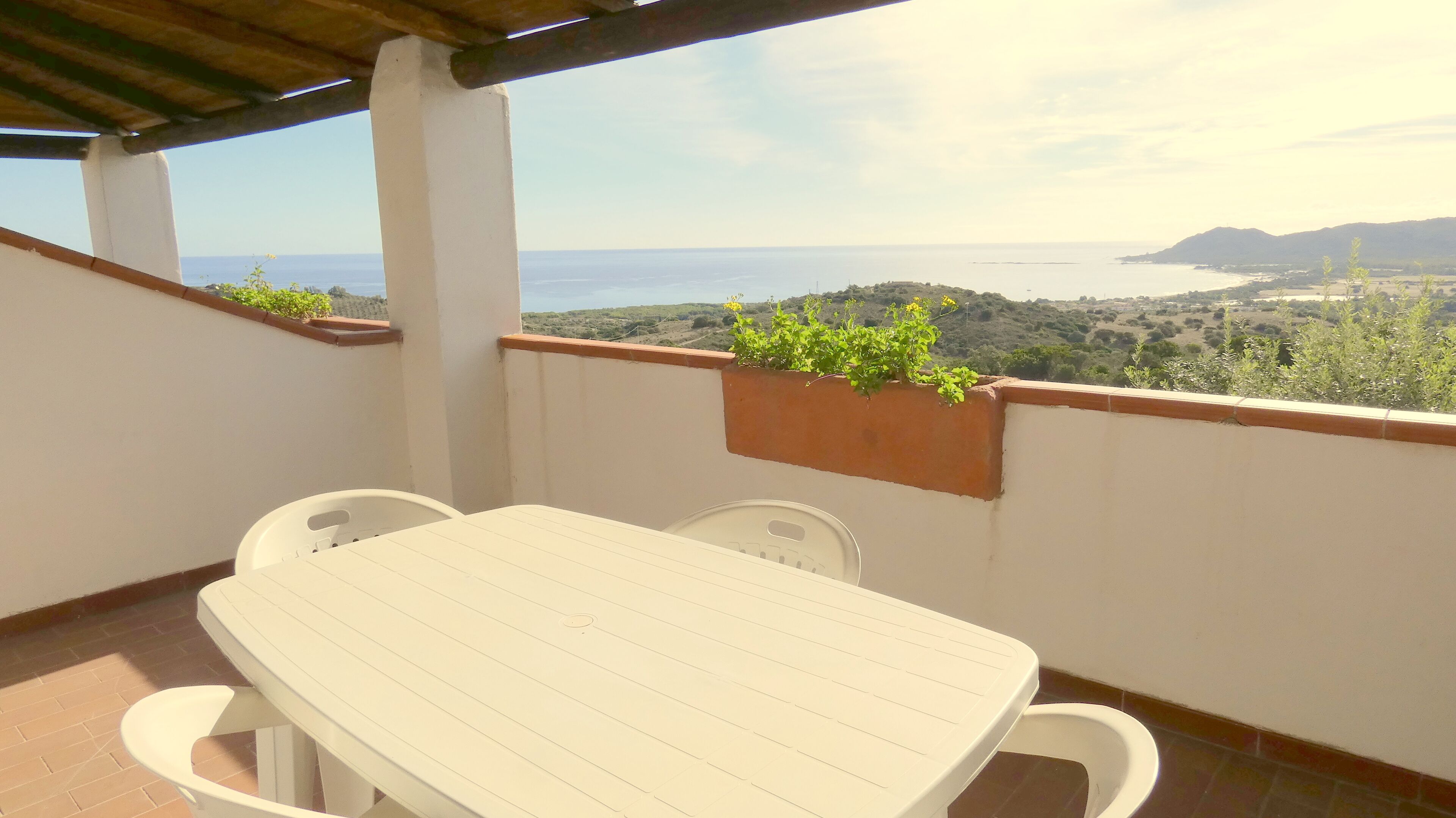 A balcony with a white table and chairs, a potted plant, and a view of the sea and hills.