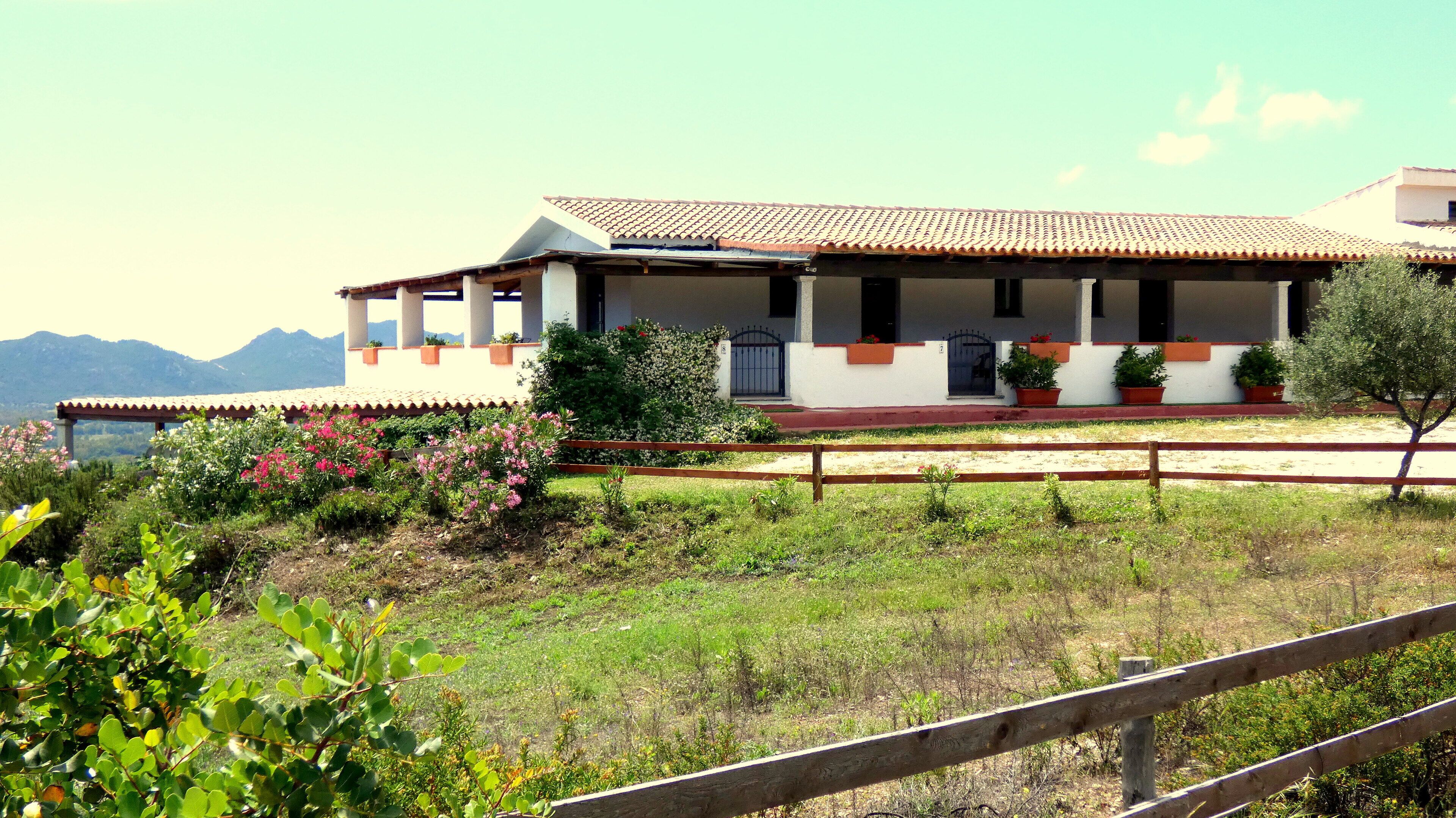A two-story building with a tiled roof, a balcony, and a garden with flowering plants.