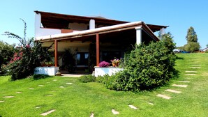 A two-story house with a covered porch, surrounded by a well-maintained lawn and various potted plants. - Comino Alto Agriturismo e  Appartamenti (Siniscola)