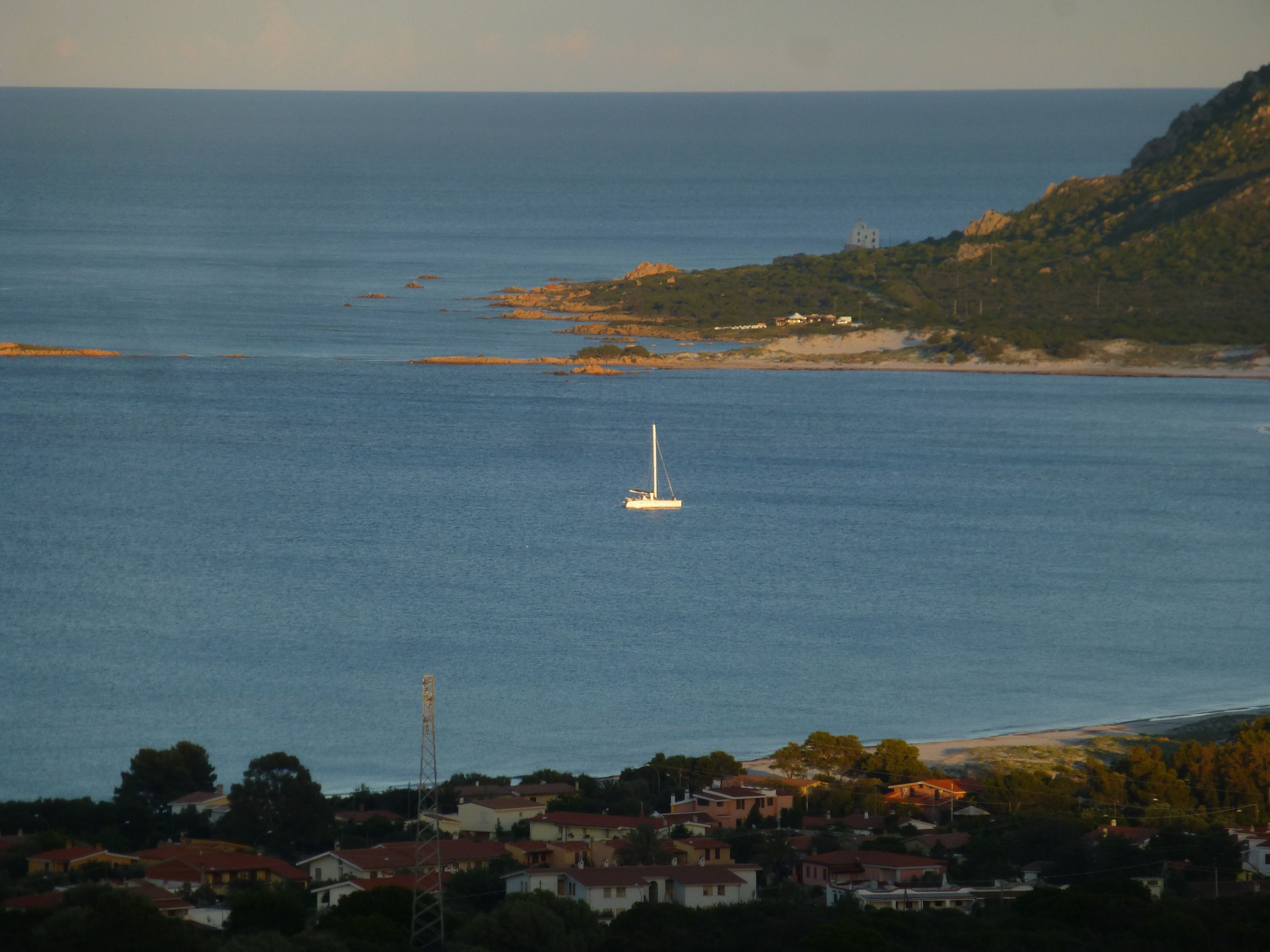 A coastal town with a sailboat on the sea, surrounded by houses and a hill with vegetation.
