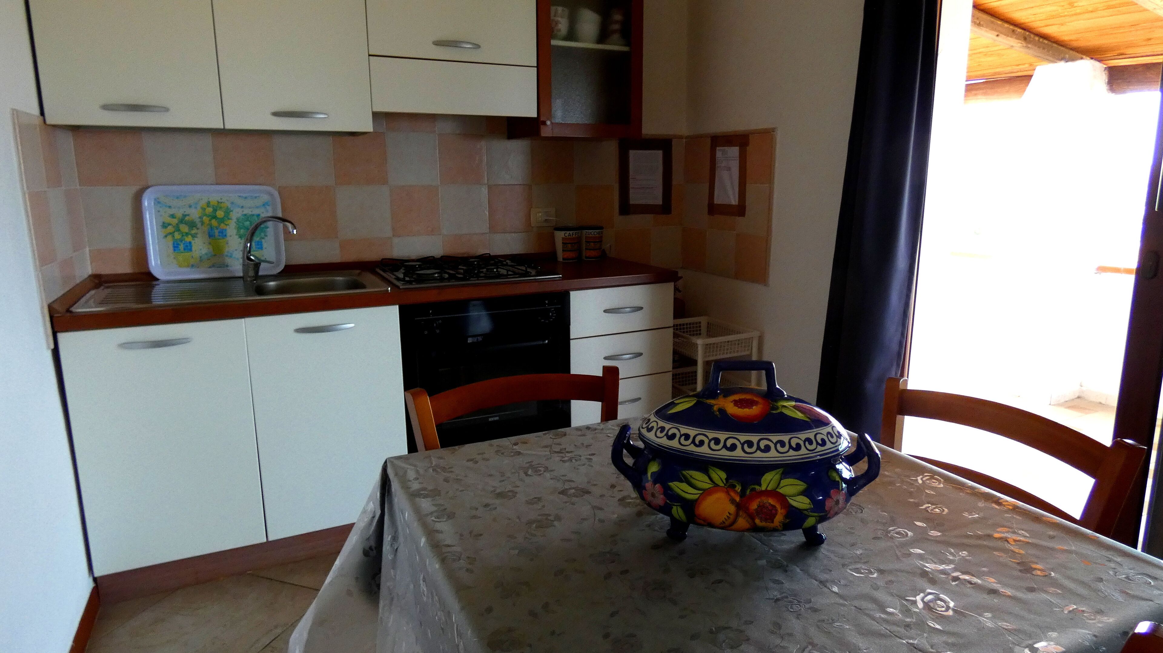 Eine kleine Küche mit weißen Schränken, einer Spüle und einem Herd. Ein Tisch mit Blumenmuster-Tischdecke und einer dekorativen Pflanze steht dort.