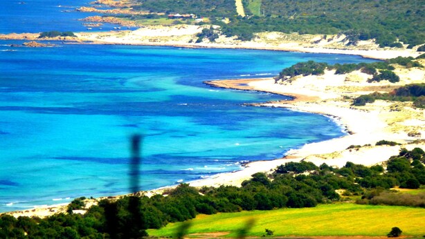 A coastal landscape with a prominent white building on a hill, a clear blue sea, and a winding road leading to the beach. - Comino Alto Agriturismo e  Appartamenti (Siniscola)