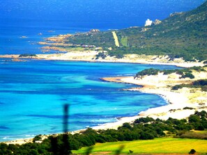 Paisagem costeira com um edifício branco imponente numa colina, mar azul límpido e uma estrada sinuosa que leva à praia.
