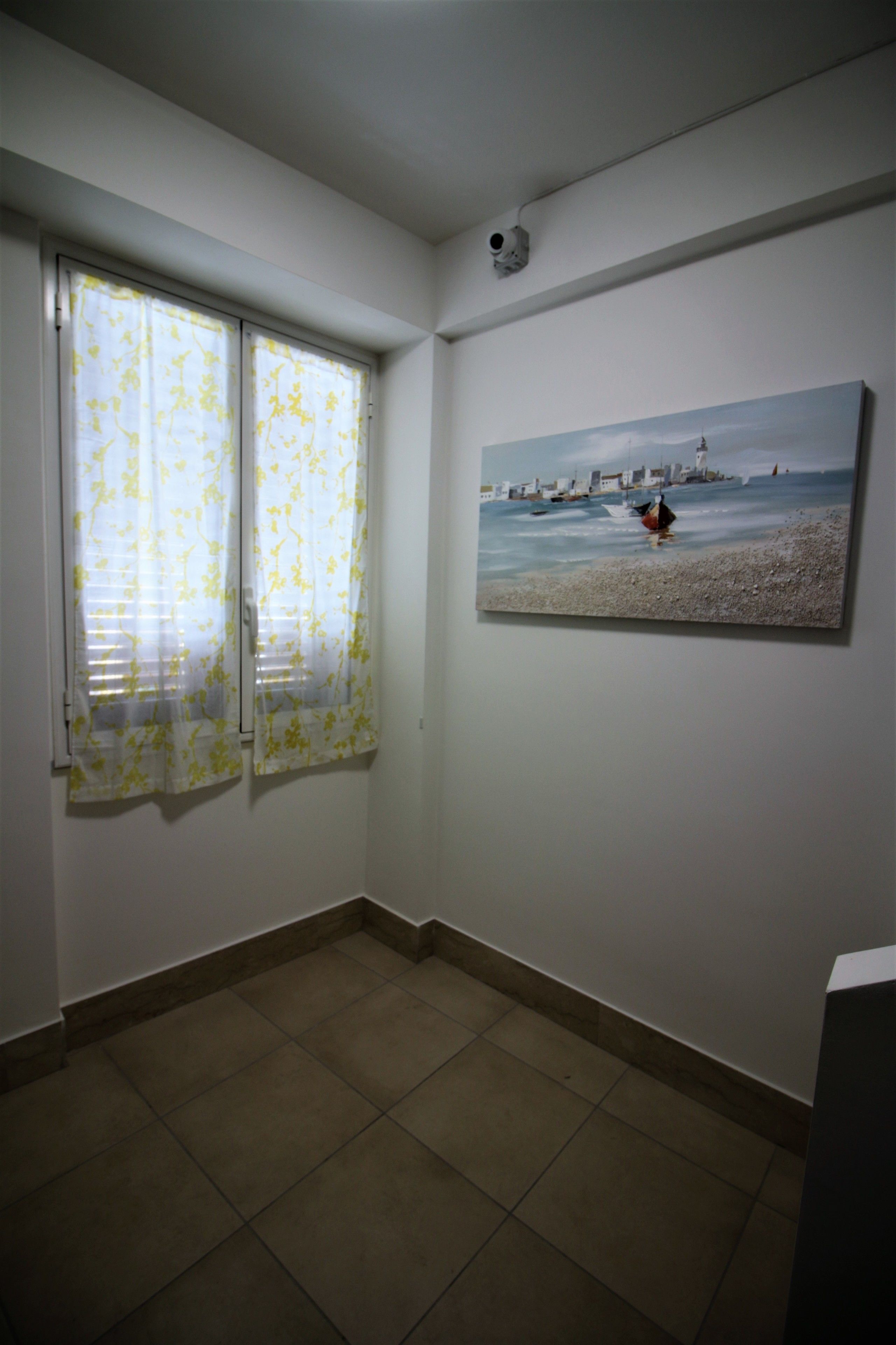 A room with a tiled floor, a painting of a seascape, and a window with floral curtains.
