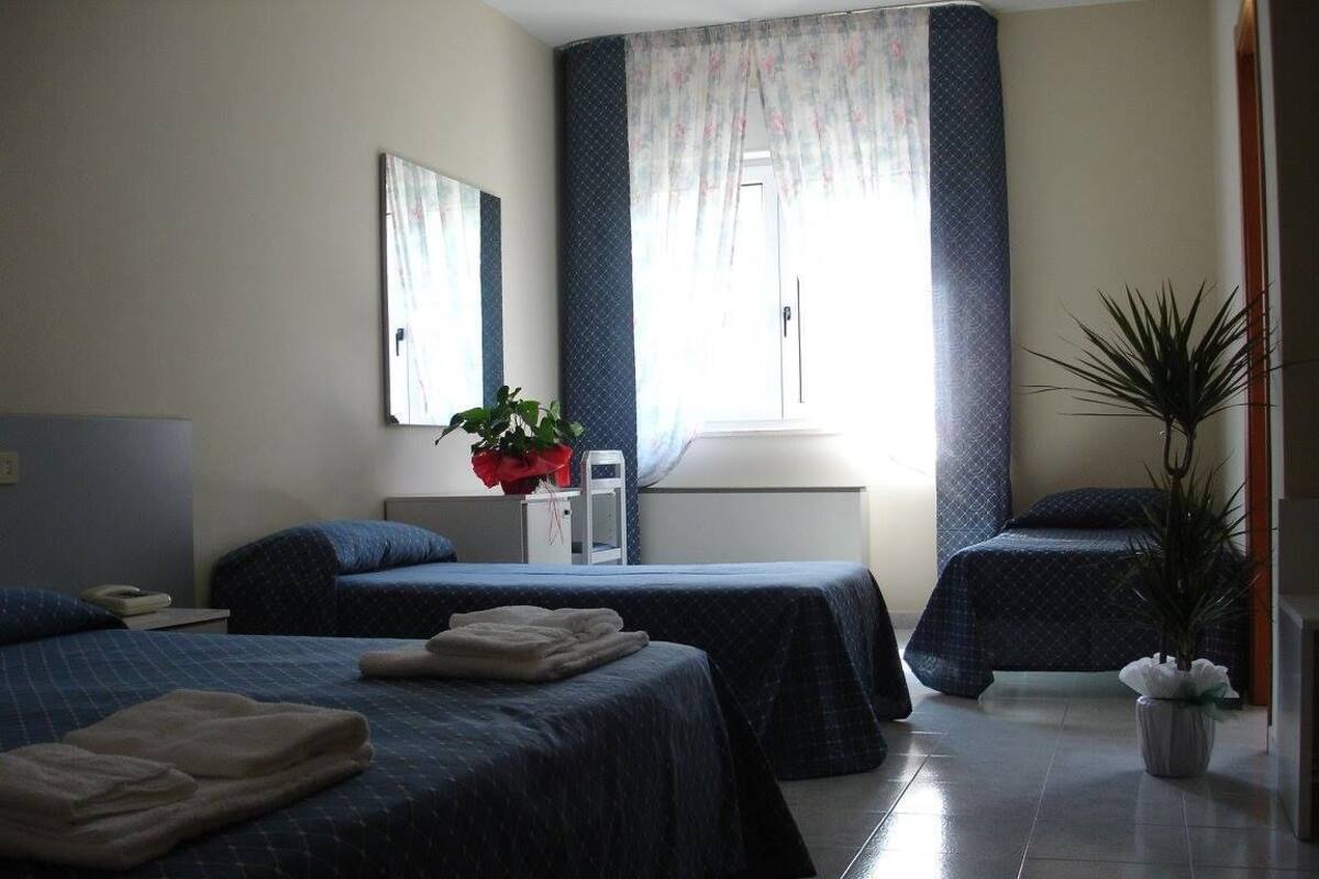 Une chambre d’hôtel avec deux lits, un petit bureau, une plante en pot et une fenêtre avec des rideaux.