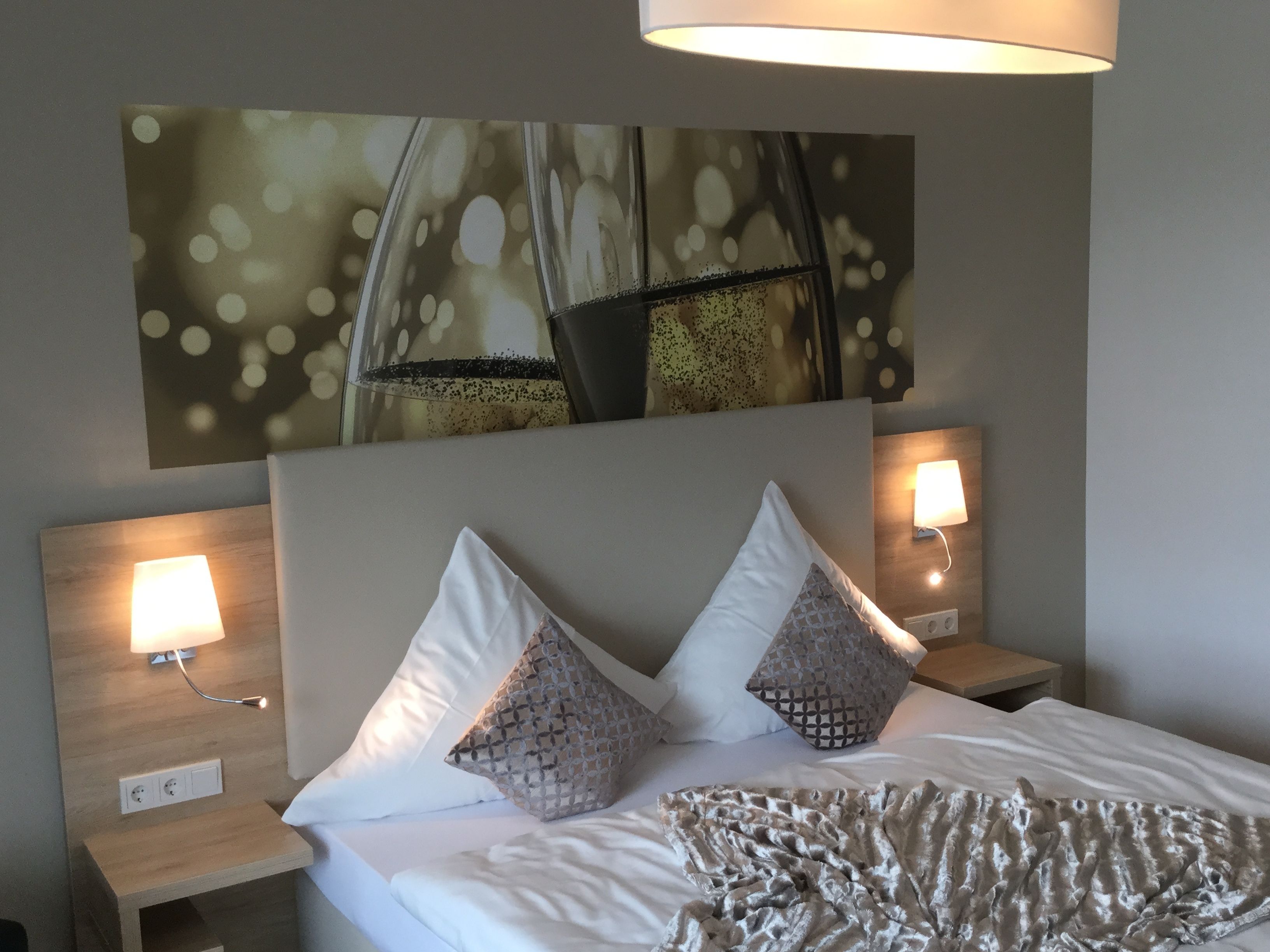 Una cama bien hecha con sábanas blancas y dos almohadas decorativas, un edredón con textura y dos lámparas de noche sobre mesitas de madera.