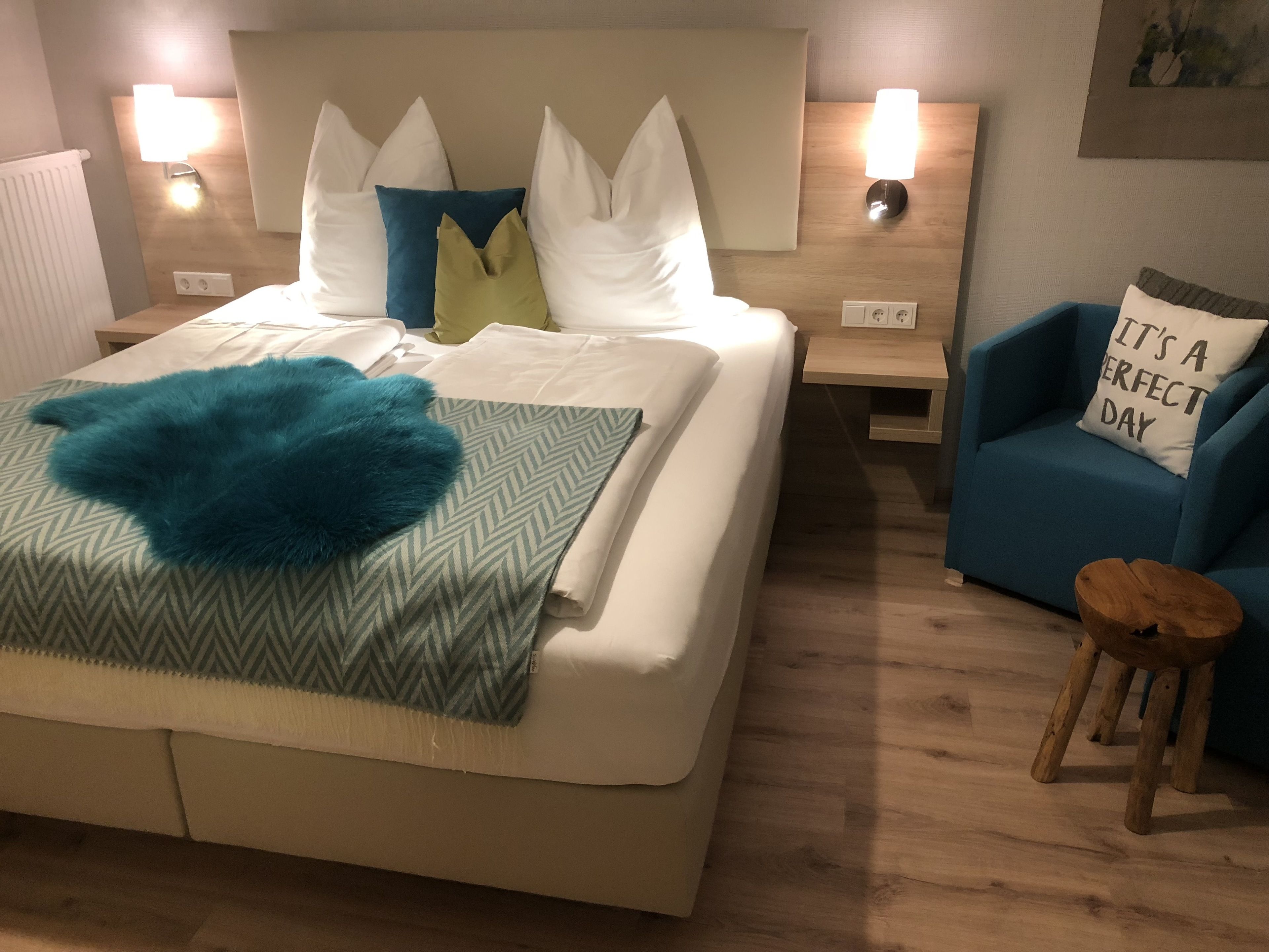 Quarto bem arrumado com cama, almofadas, uma cadeira azul, um banquinho de madeira e uma mesa de cabeceira com abajur.
