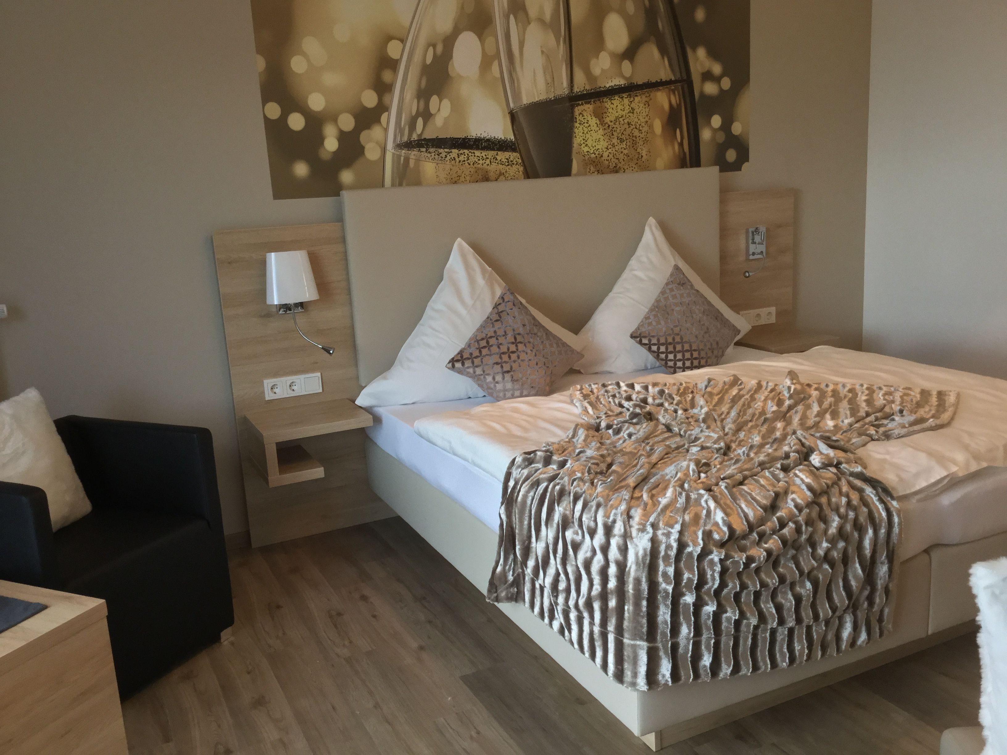 A modern bedroom with a large bed, a bedside table, a nightstand, a lamp, and a wall-mounted shelf.
