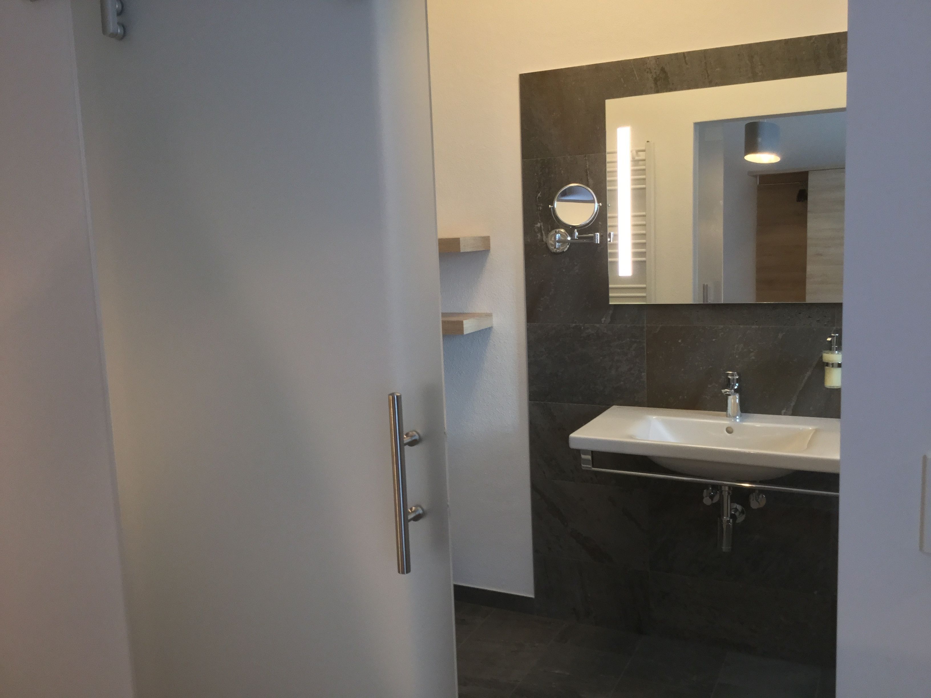 Een badkamer met een wit wastafelmeubel, donkere tegels, een spiegel en een deur.