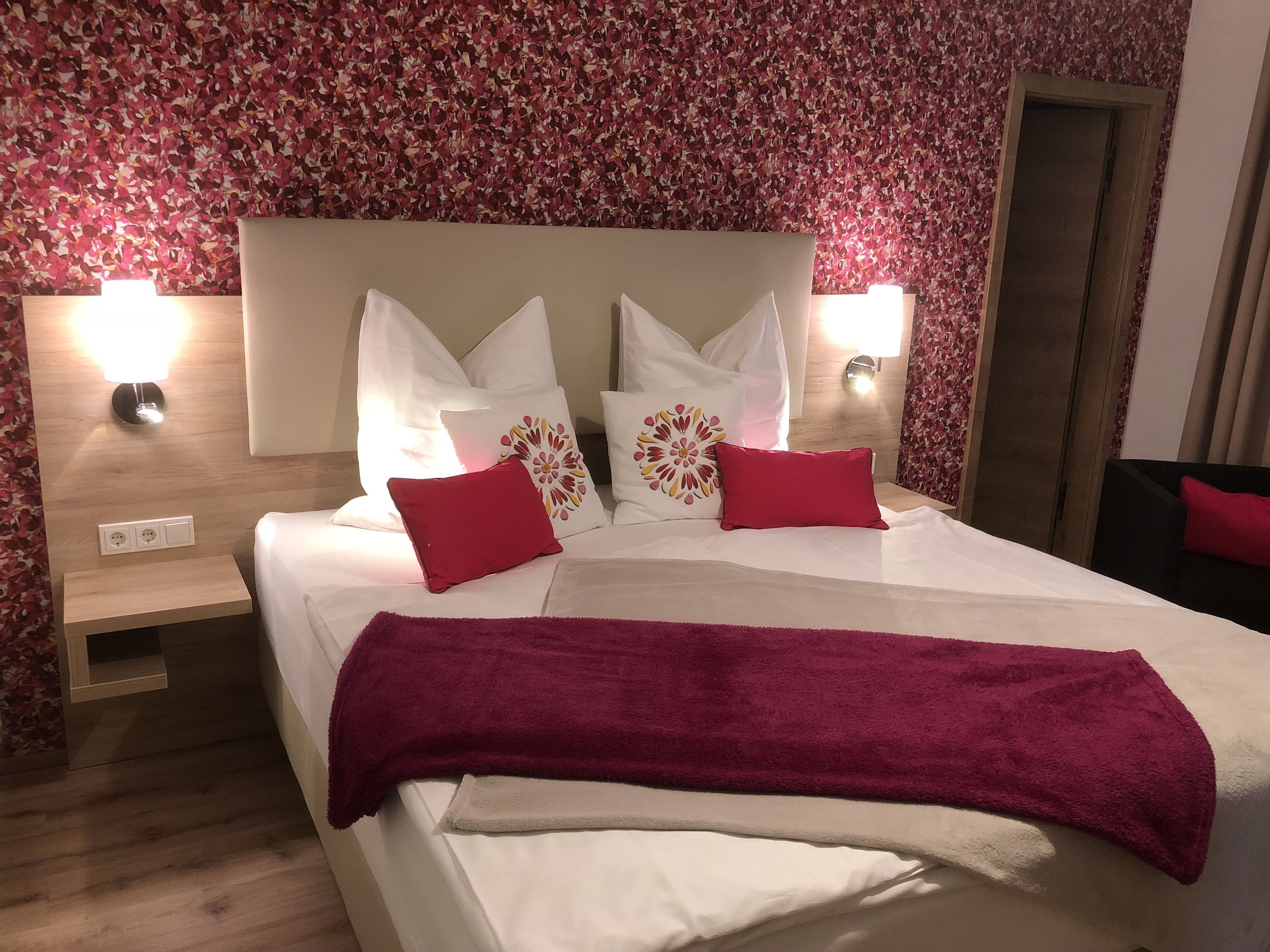 A neatly made bed with white linens, two red pillows, and a purple throw blanket. There are two wall-mounted lamps on either side of the bed.