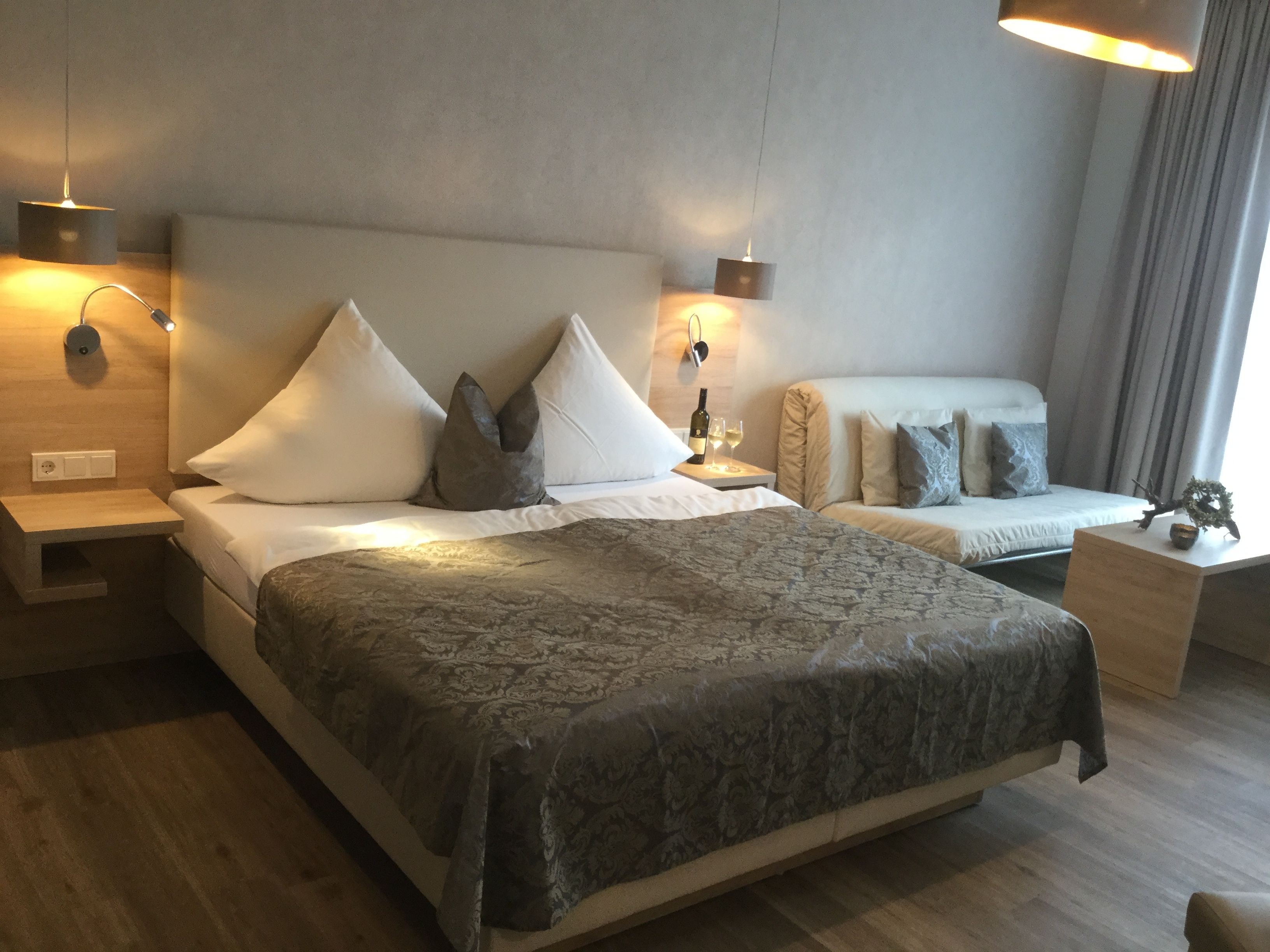 A modern hotel room with a large bed, two bedside tables, a nightstand, and a small table with a bottle and glass.