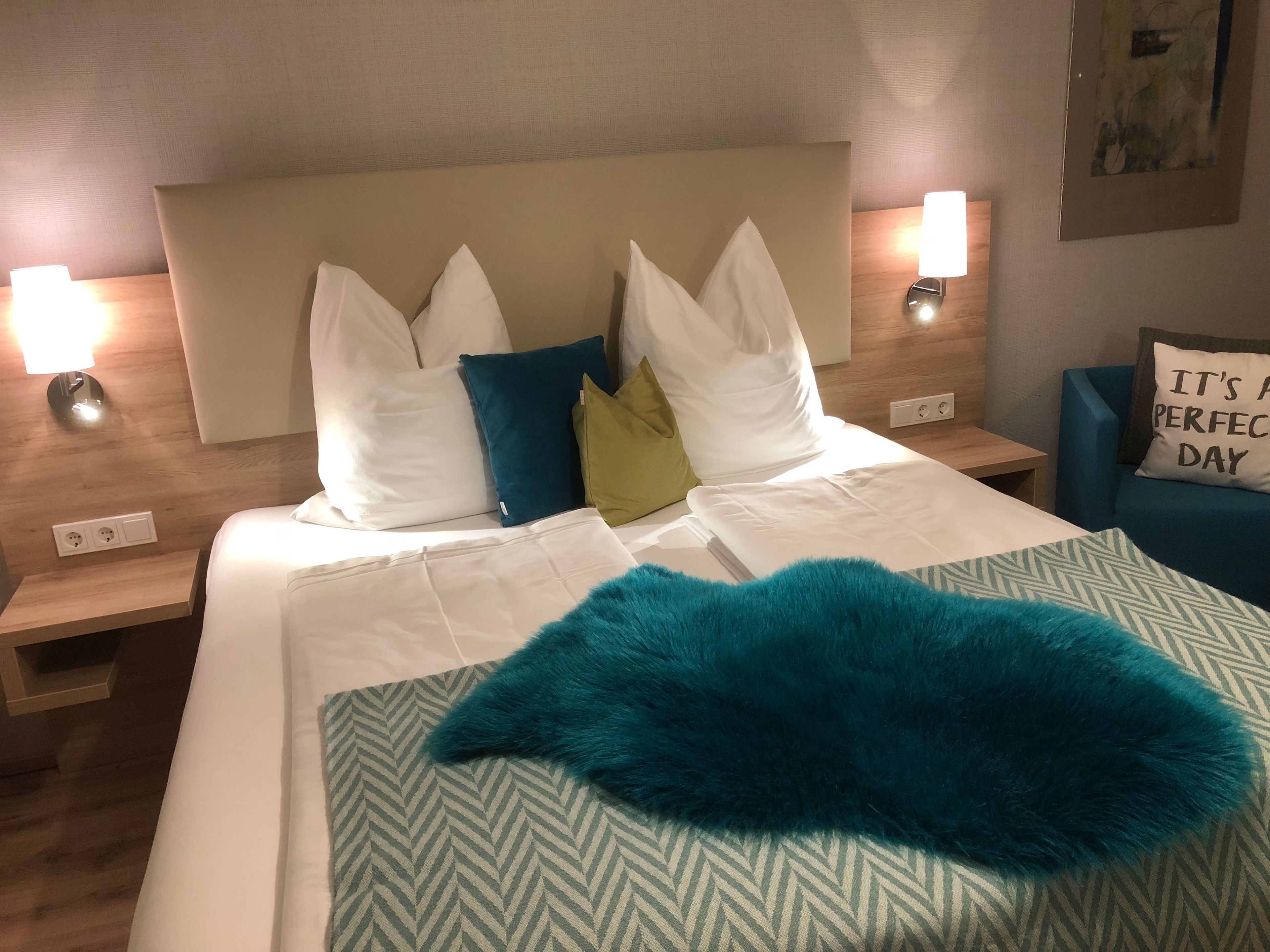 A neatly made bed with white linens, a teal throw blanket, and a variety of pillows in white, blue, and green. There are two bedside lamps on wooden nightstands.