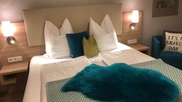 A neatly made bed with white linens, a teal throw blanket, and a variety of pillows in white, blue, and green. There are two bedside lamps on wooden nightstands.