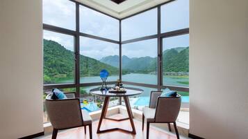 A room with a large window overlooking a lake and mountains, a round table with chairs, and a small pool.
