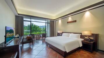 A hotel room with a large bed, a desk with a flat-screen TV, a balcony with a view of greenery, and a decorative wall piece.