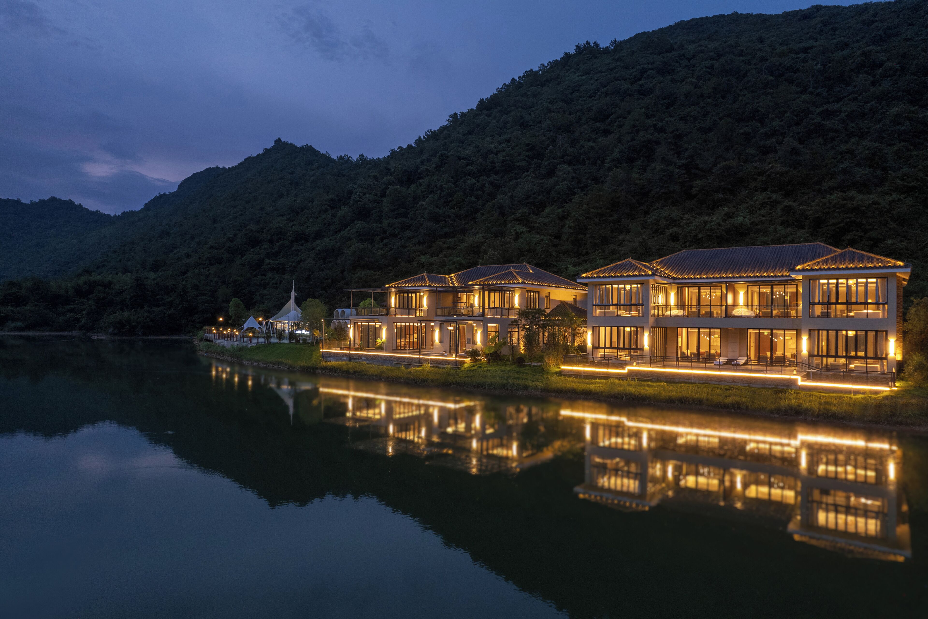 A waterfront property with multiple buildings, a reflective lake, and a forested hill in the background.