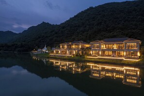 Una propiedad frente al agua con varios edificios, un lago que se refleja y una colina boscosa al fondo.