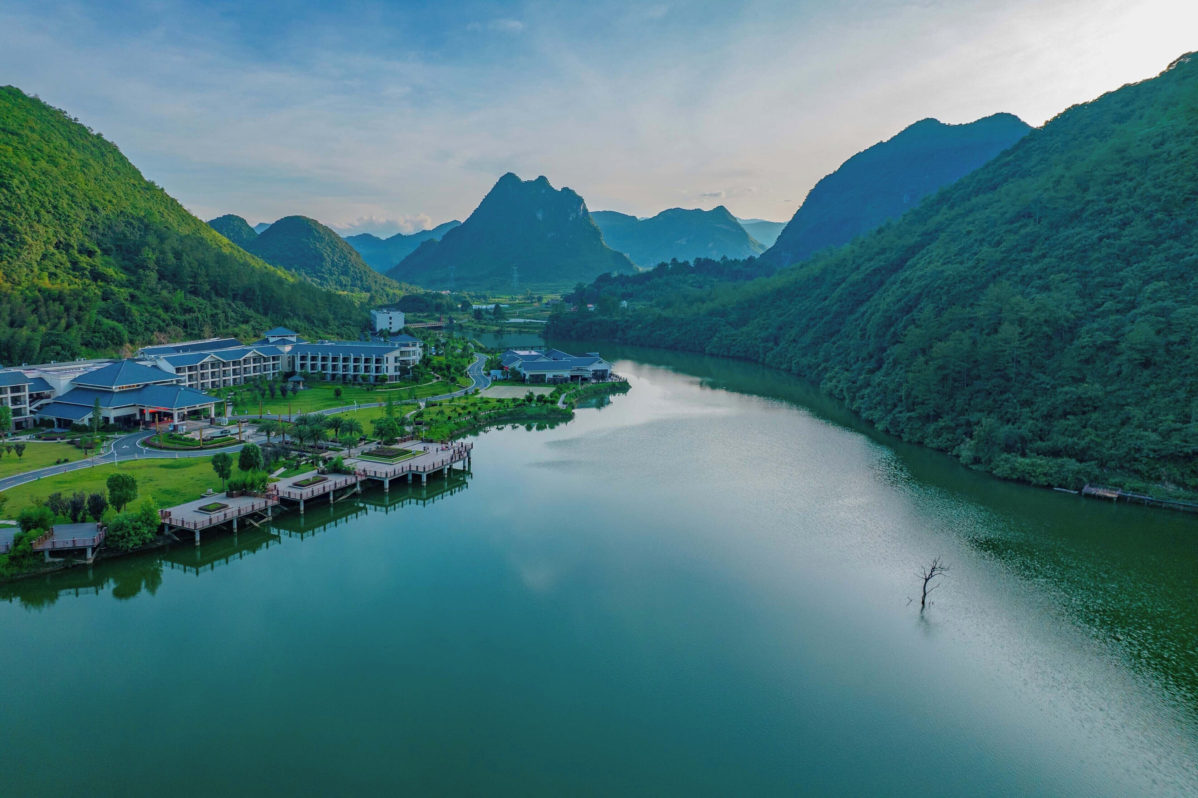 A riverside hotel with a dock, surrounded by lush green hills and a mountain in the background.