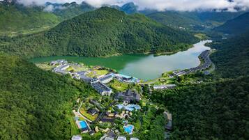 A resort with multiple buildings, a swimming pool, and a lake surrounded by forested hills.