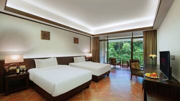 A hotel room with two beds, a desk, a TV, and a balcony with a view of trees.