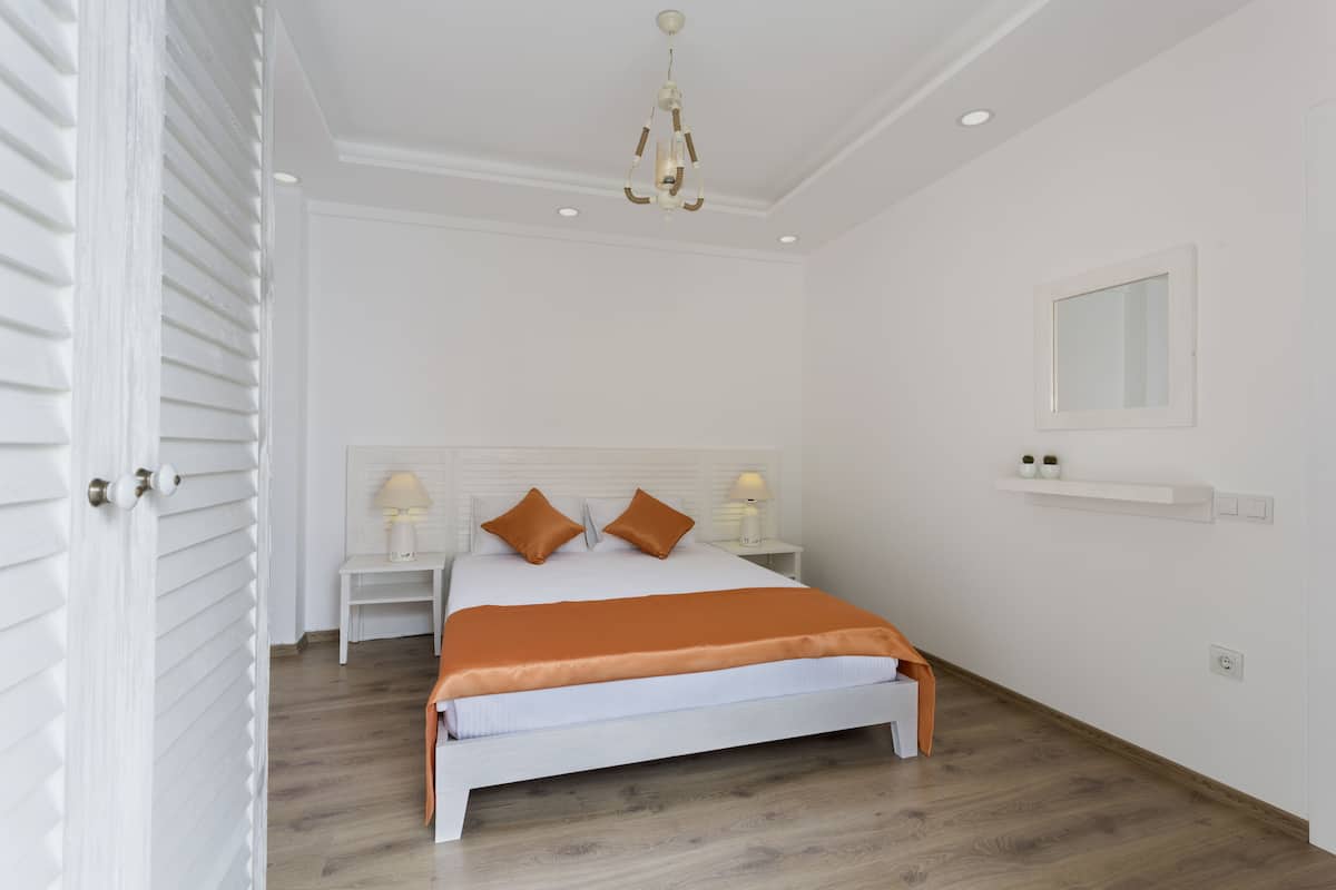 A bedroom with a white bed, two pillows, a nightstand with a lamp, a ceiling light, and a small shelf with decorative items.