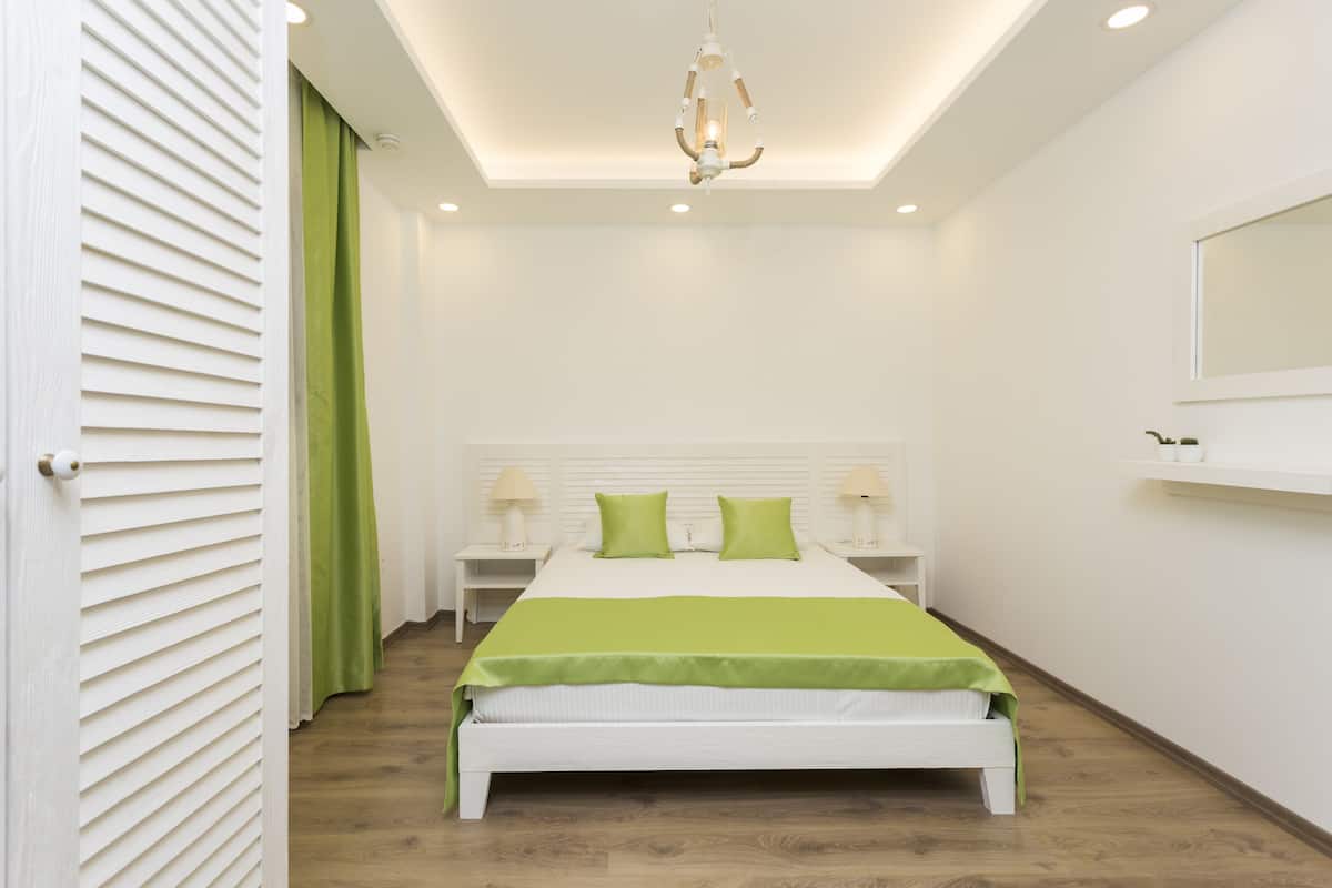 A modern bedroom with a white bed, green pillows, and a green throw blanket. There are two bedside tables with lamps, a ceiling light, and a window with a white frame.