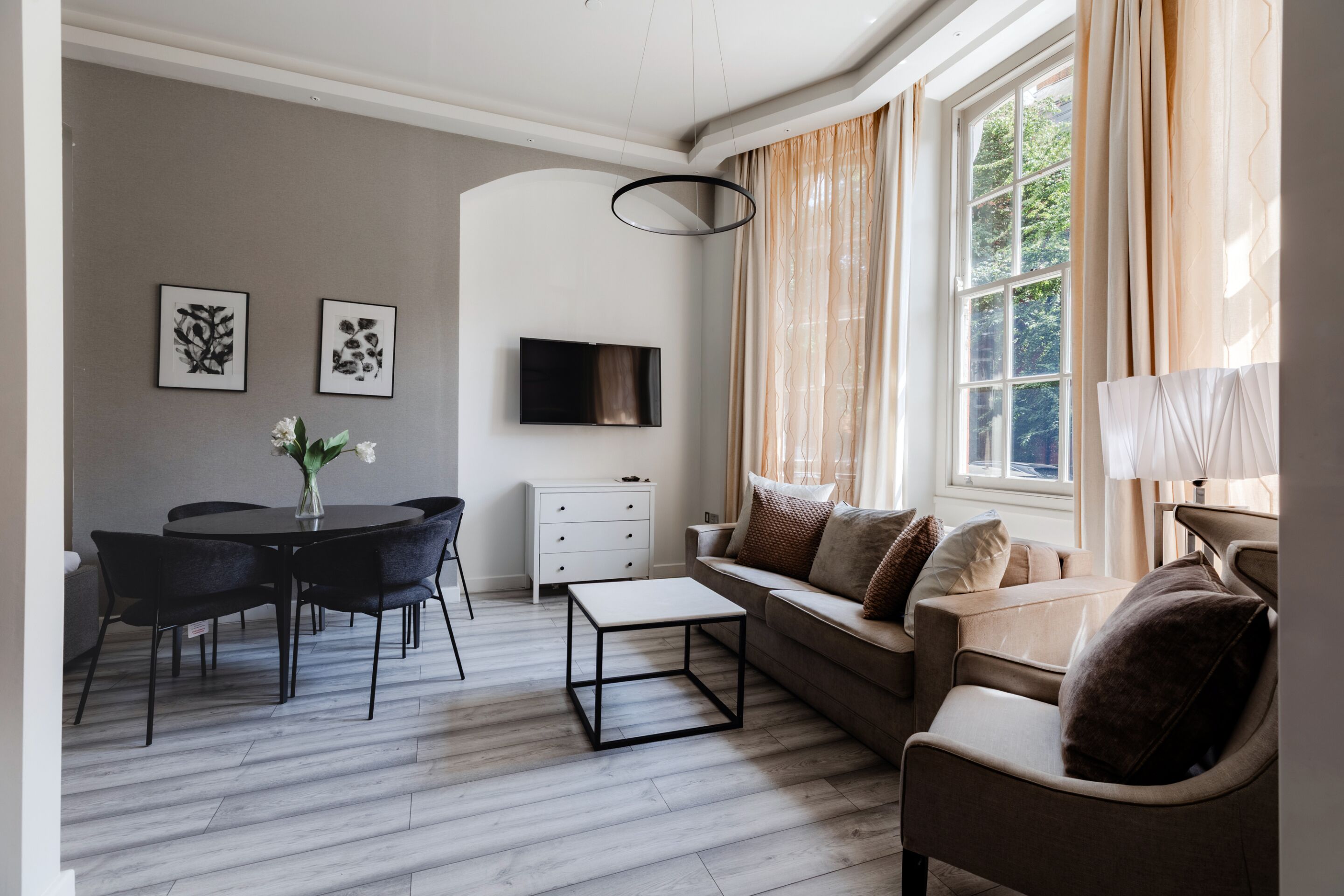 A modern living room with a sofa, a coffee table, a dining table with chairs, a flat-screen TV mounted on the wall, and a window with curtains.
