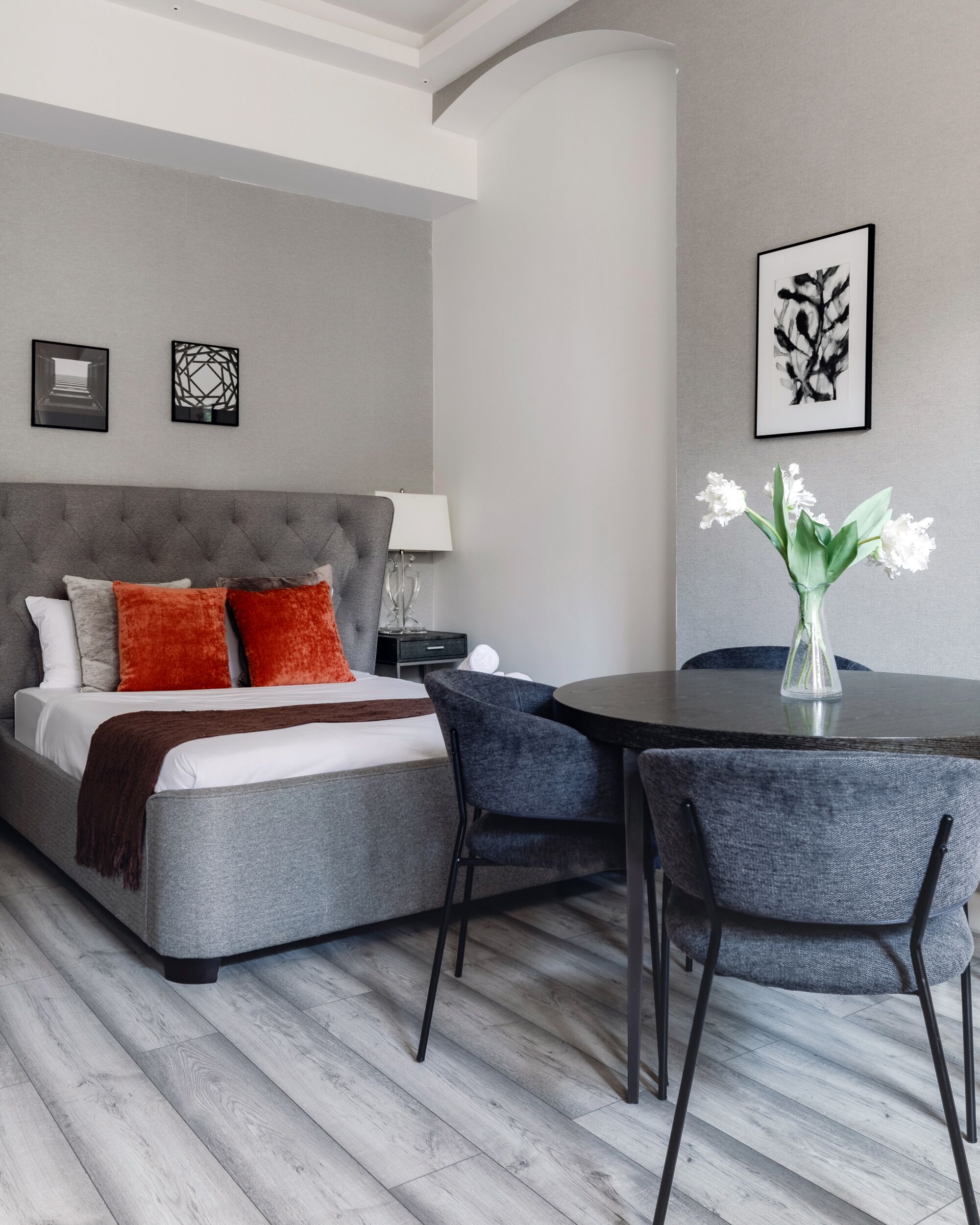 A modern hotel room with a grey upholstered bed, a small round table with two chairs, a vase with white flowers, and two framed artworks on the wall.