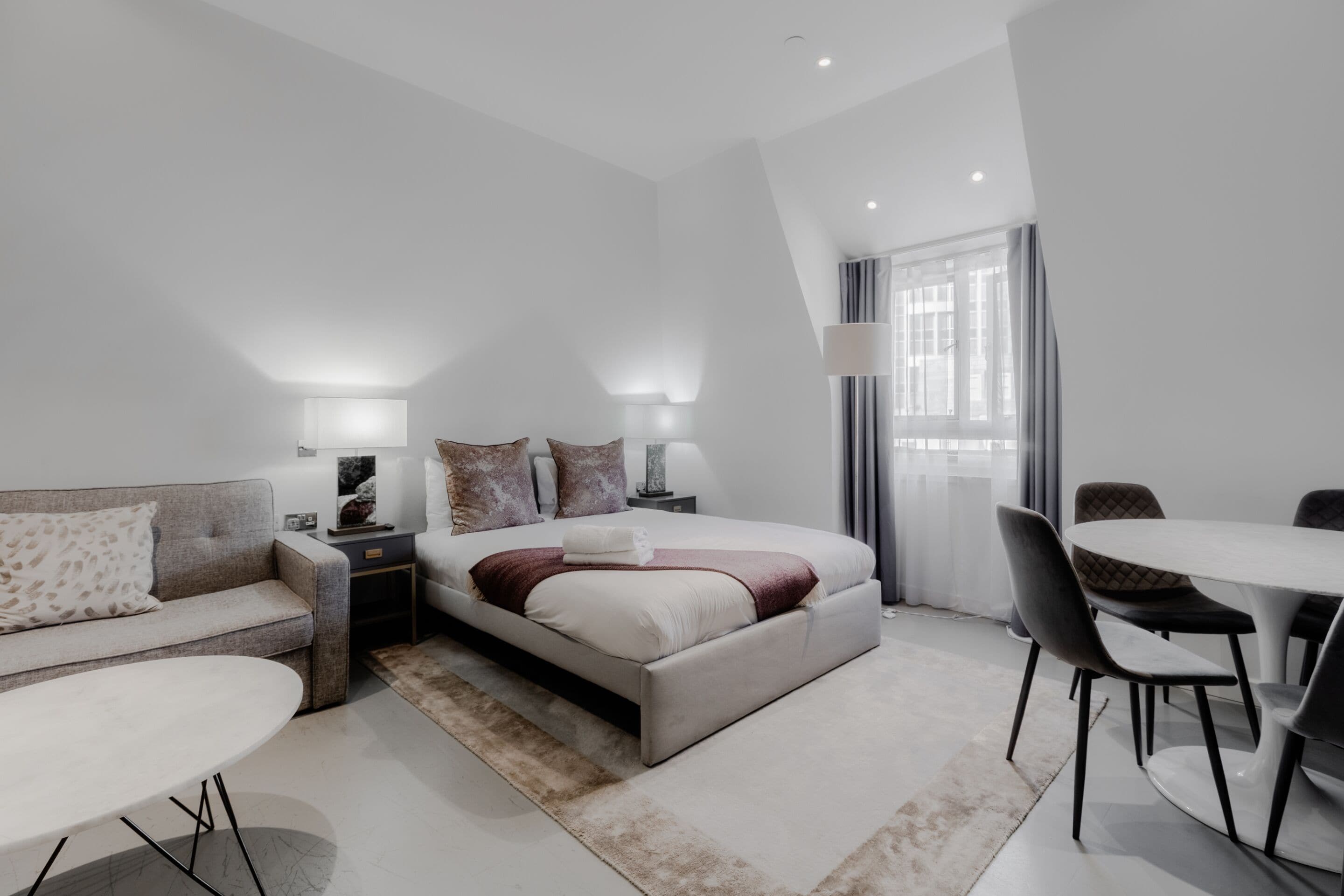 A modern hotel room with a large bed, a sofa, a dining table, and a lamp.