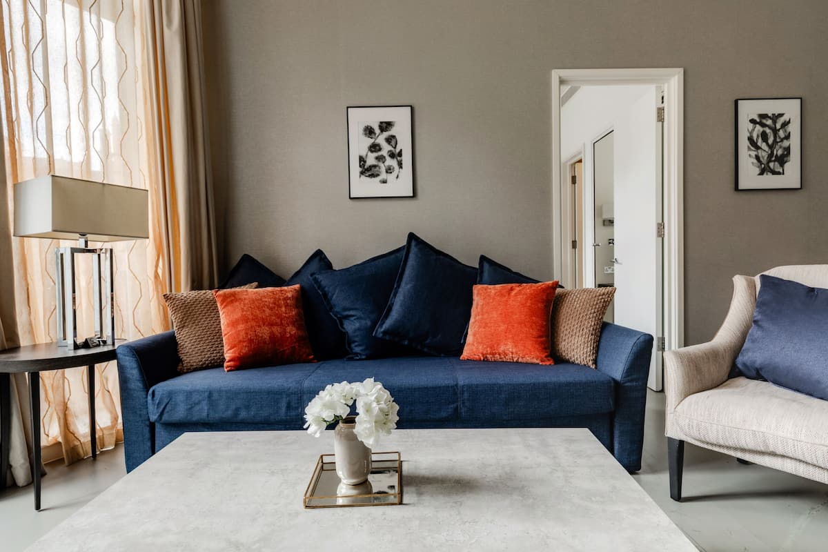 Un salon moderne avec un canapé bleu, des coussins orange et marron, un vase blanc orné de fleurs et deux œuvres d’art encadrées accrochées au mur.