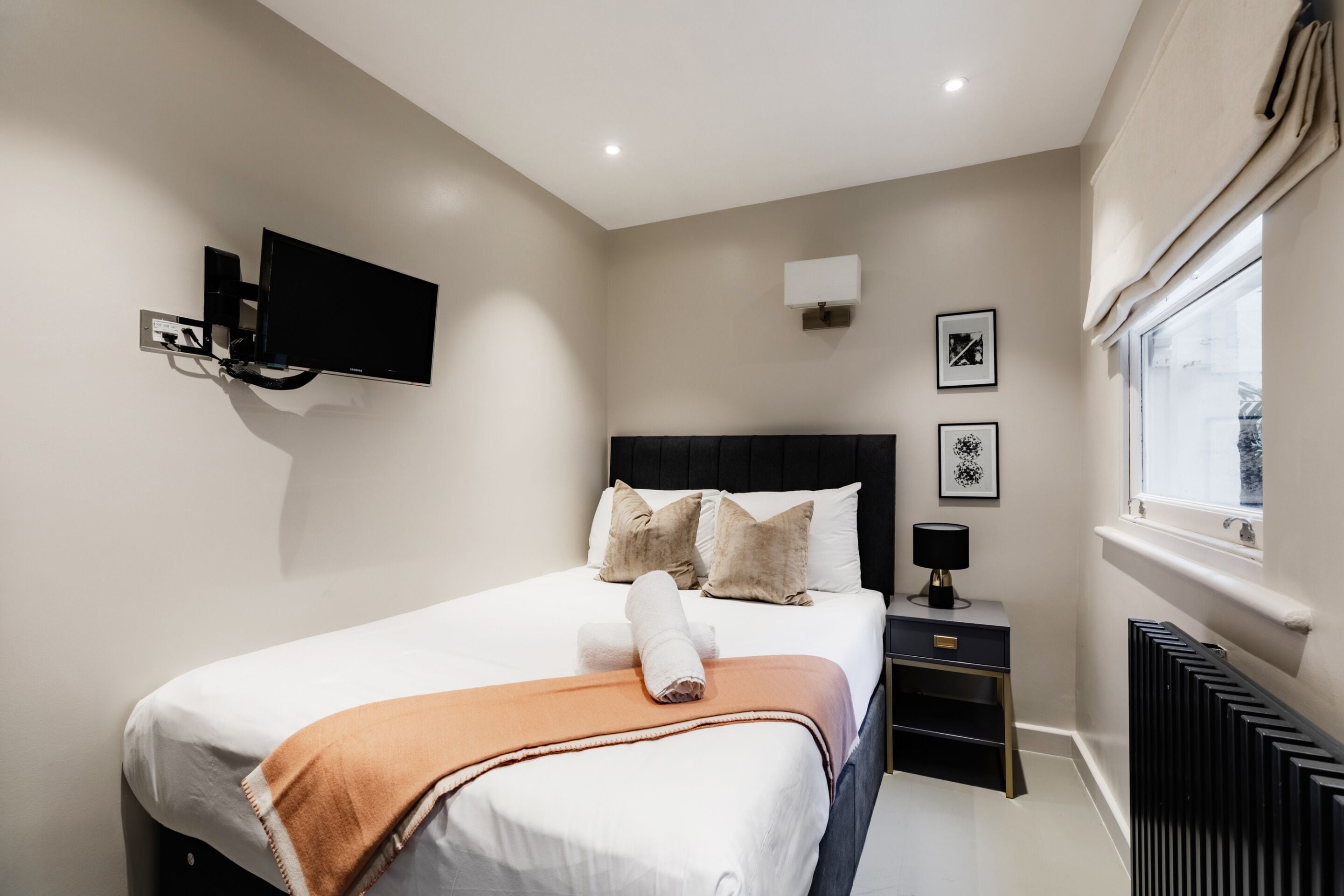 A modern hotel room with a large bed, a flat-screen TV mounted on the wall, a bedside table with a lamp, and a window with a view.