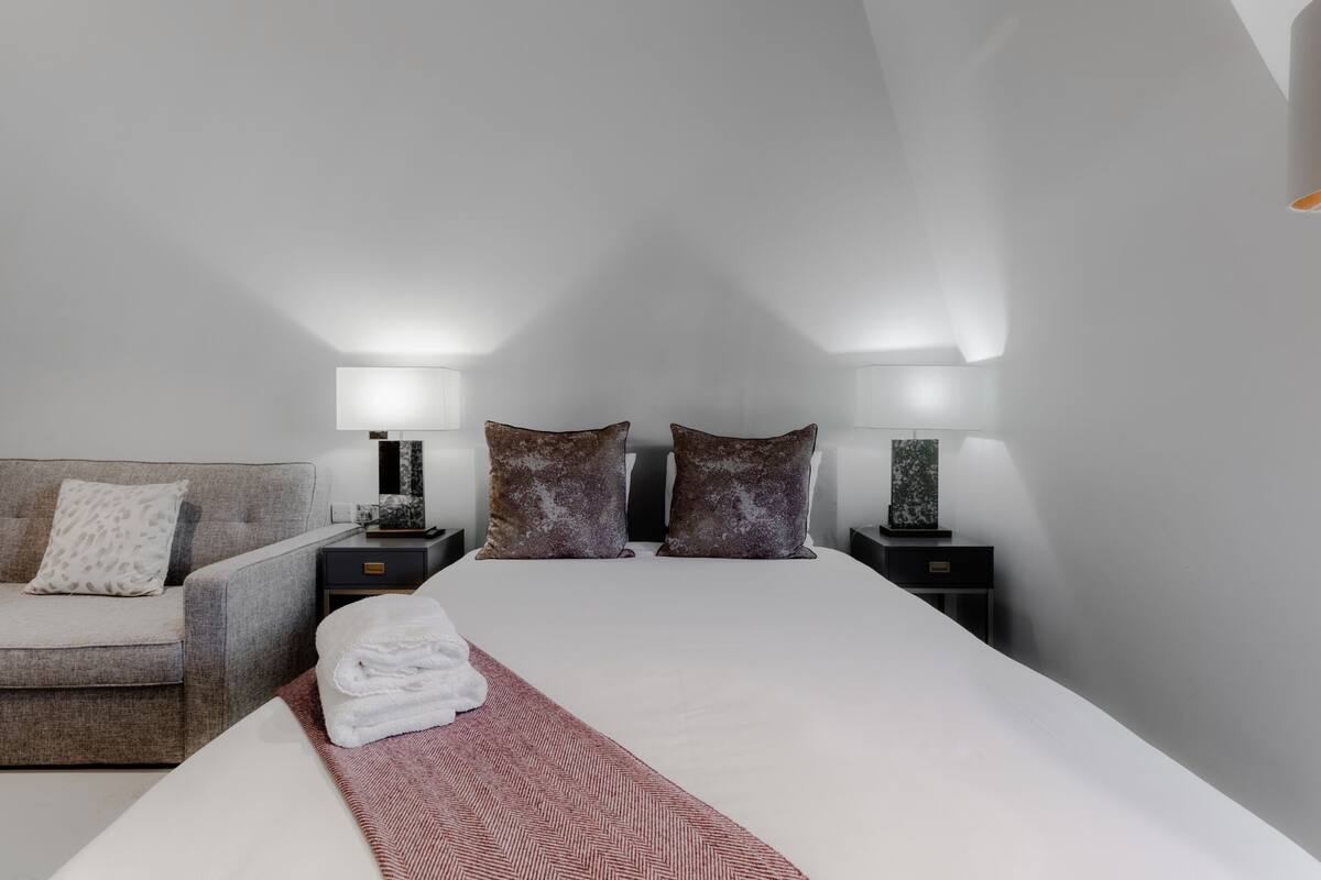 Une chambre d’hôtel moderne avec un lit soigneusement fait, un canapé, deux tables de chevet avec des lampes et un tapis décoré d’un motif en forme d’arête de hareng.