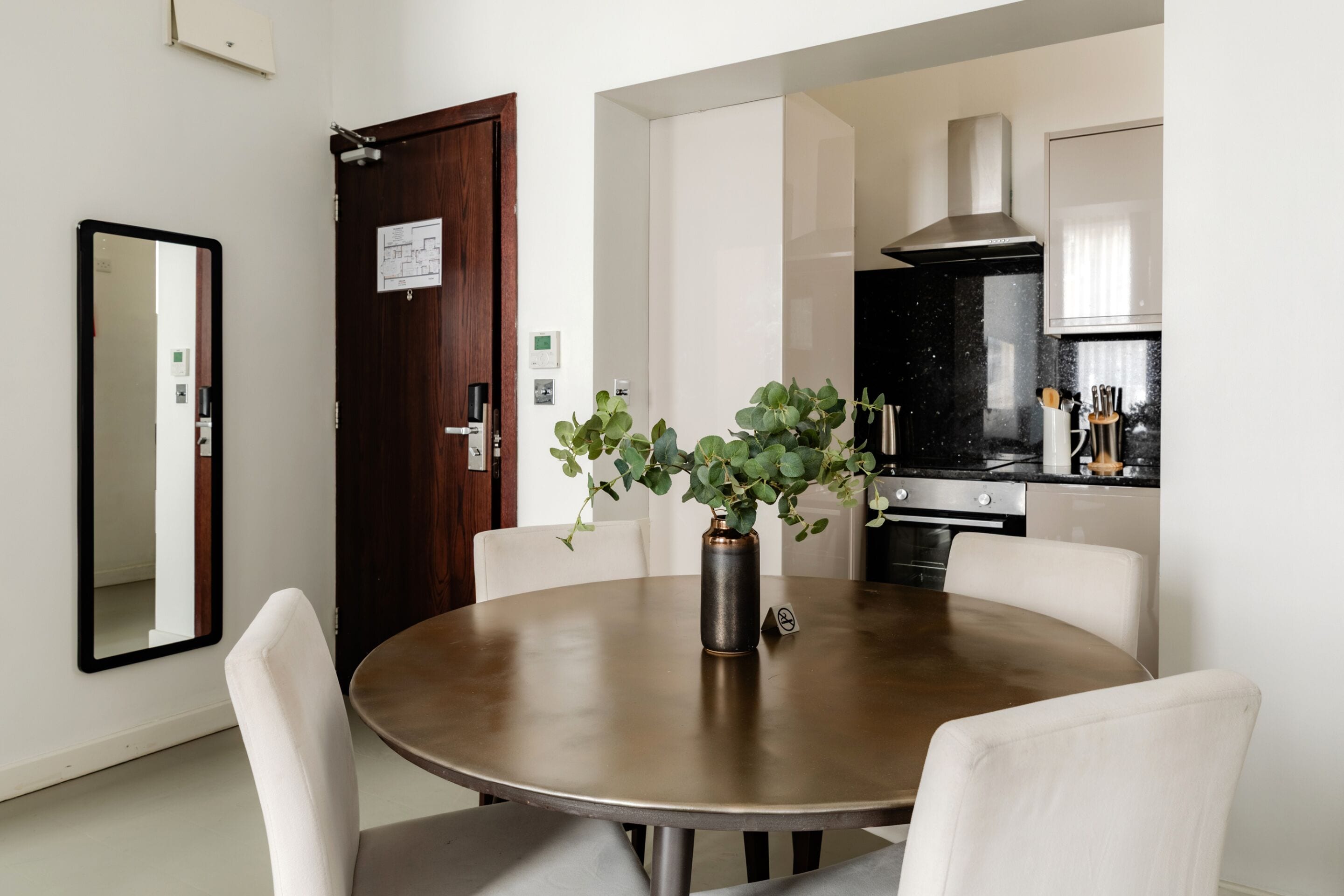 Un área de comedor con una mesa redonda, sillas blancas, un florero con plantas verdes, una puerta de madera y una cocina al fondo.