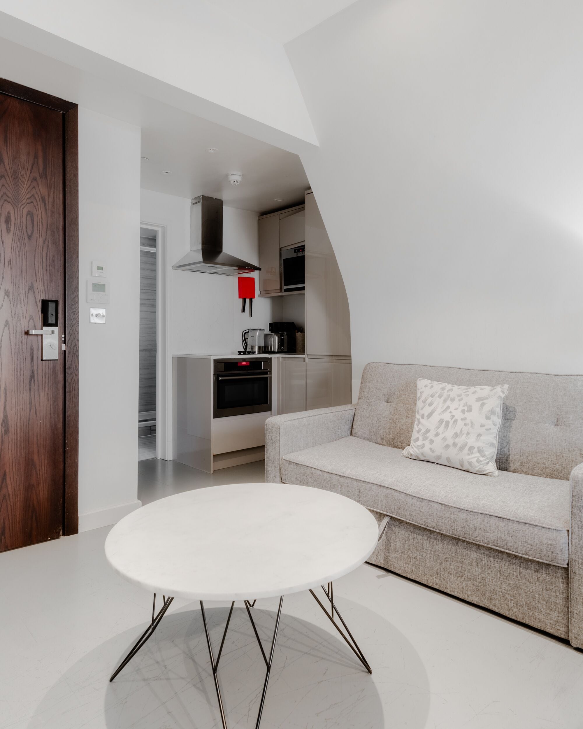 Uma sala de estar moderna com uma mesa redonda branca, um sofá cinza com almofada estampada e uma porta de madeira.