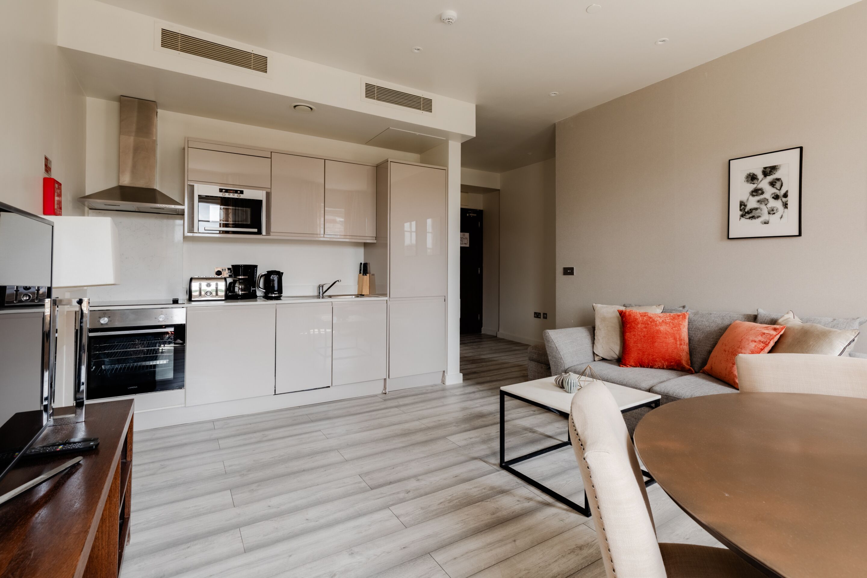 A modern kitchen with white cabinetry, a built-in oven, and a coffee maker. There is a dining area with a wooden table and chairs.