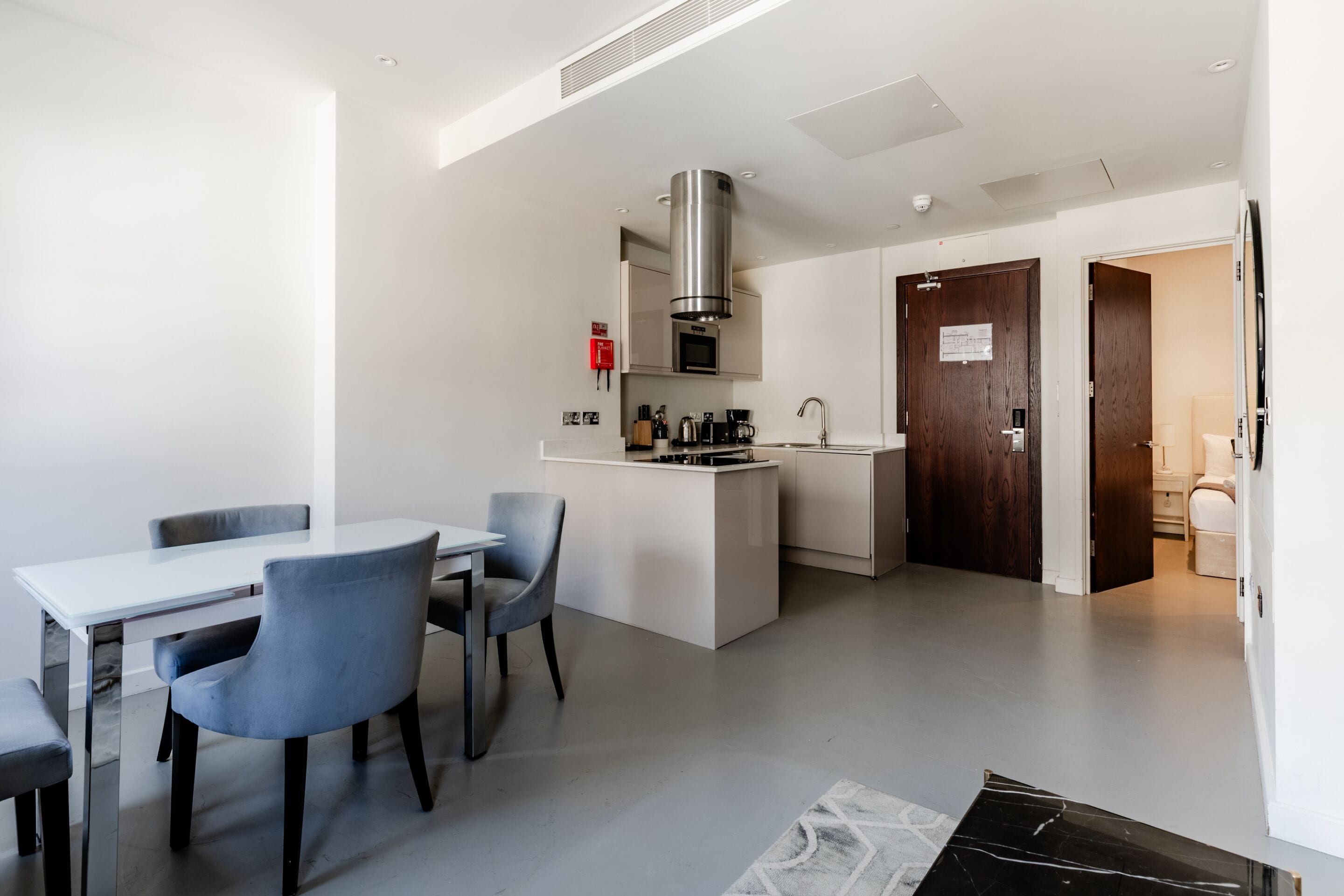 A modern kitchen with a central island, a dining area with a white table and grey chairs, and a dark wooden door leading to another room.