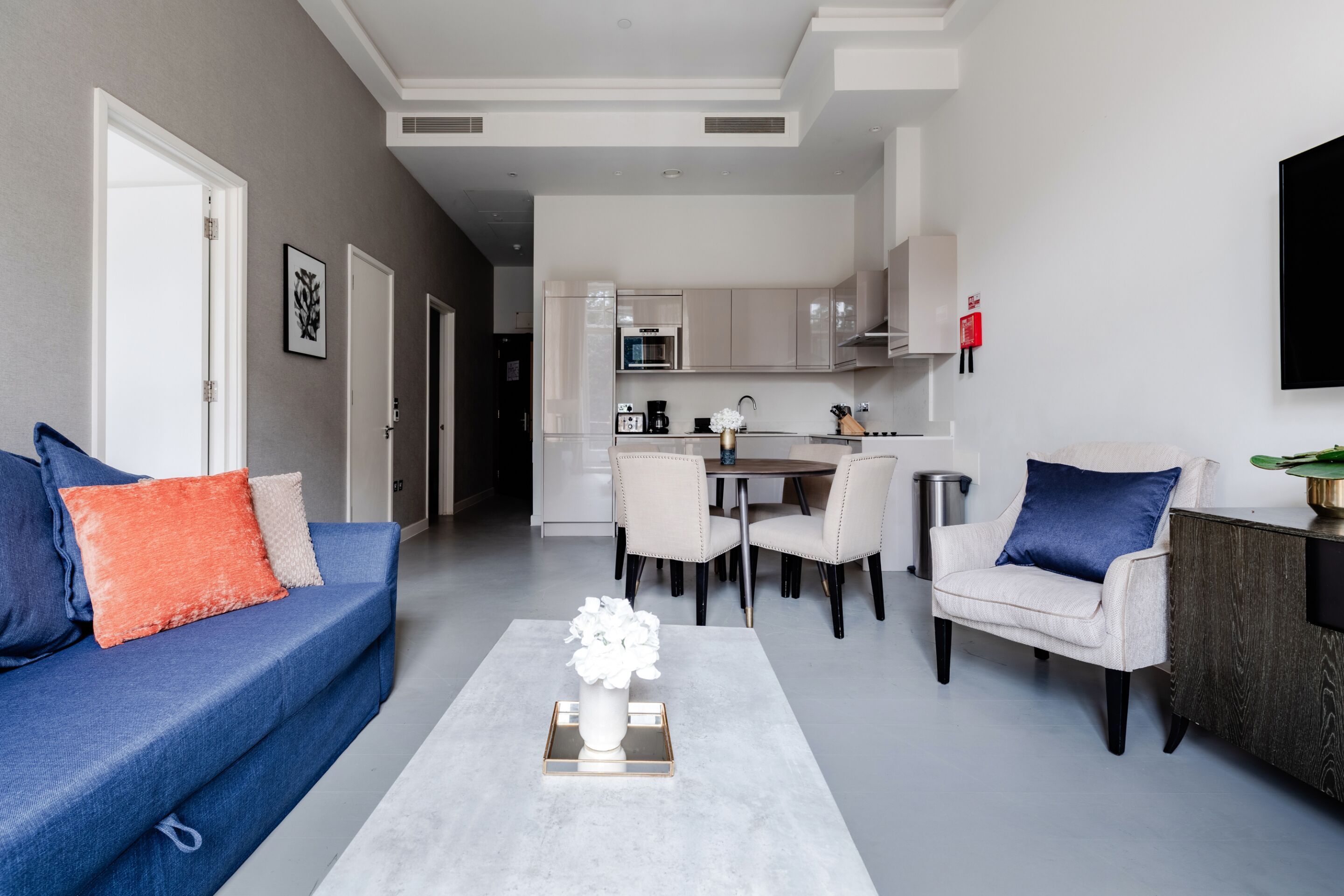 Uma sala de estar moderna com sofá, poltronas, mesa de centro e área de jantar, com uma cozinha ao fundo.