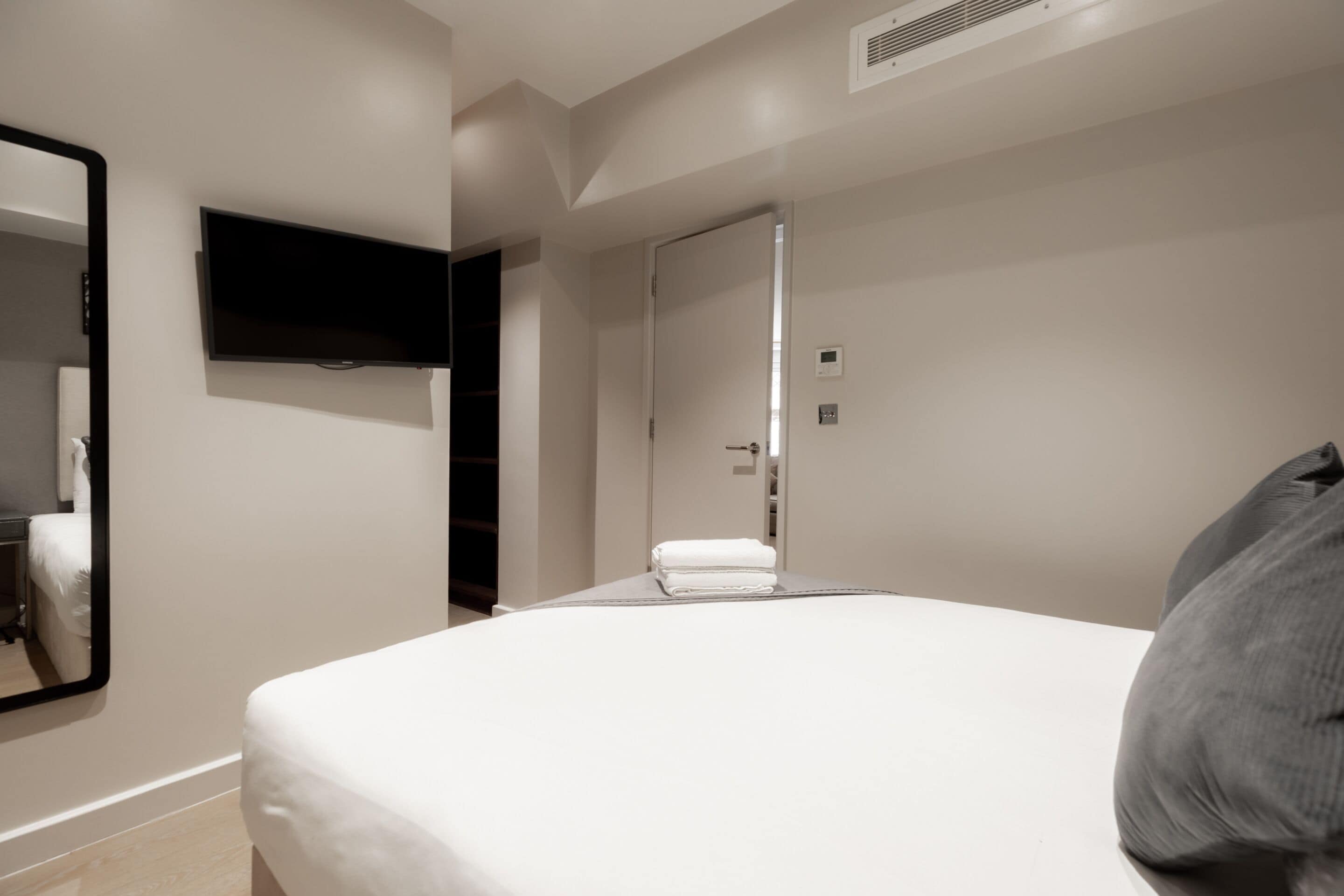 A modern hotel room with a large bed, a flat-screen TV mounted on the wall, a mirror, and a door leading to a bathroom.
