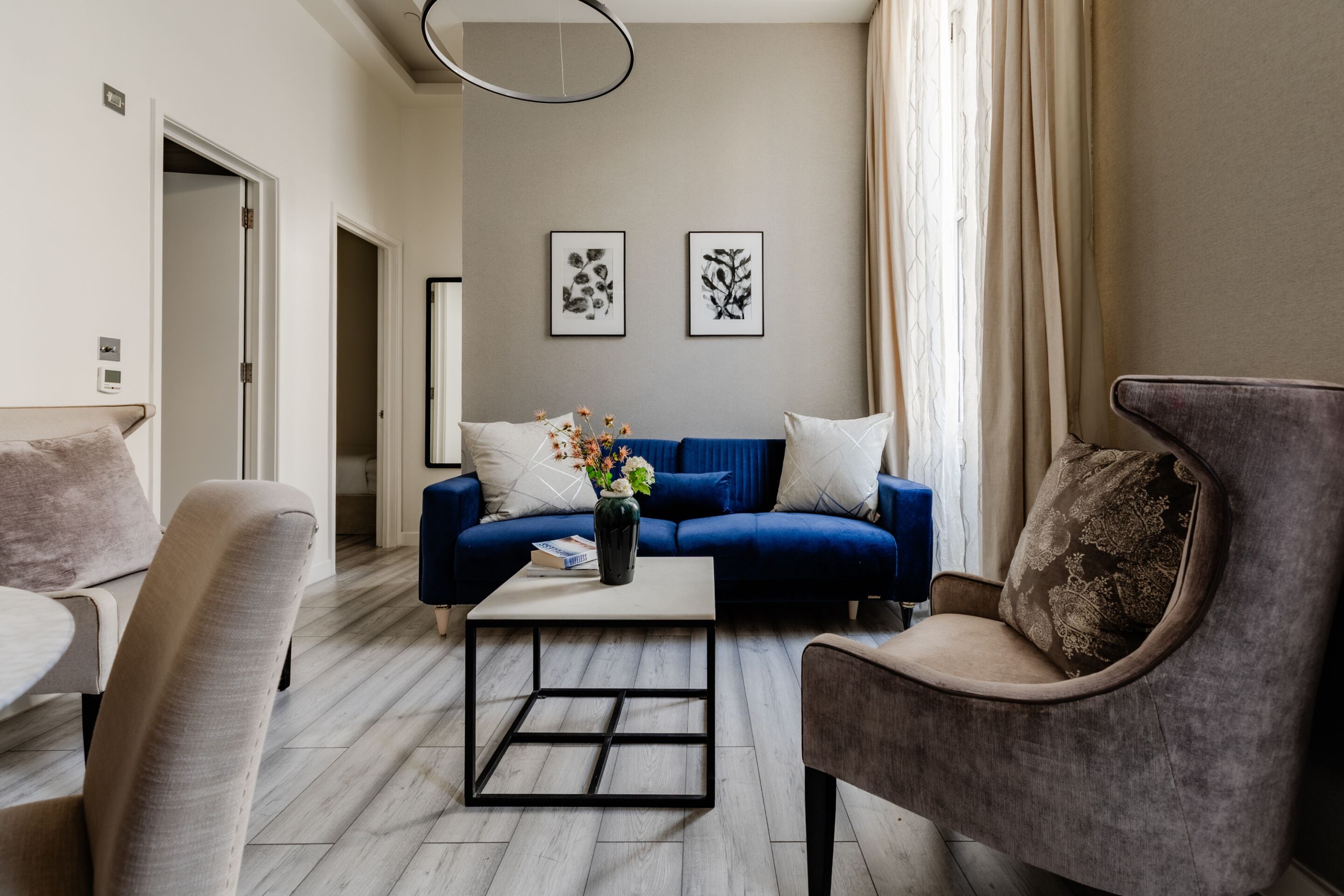 Een moderne woonkamer met een blauwe bank, een salontafel en een grijze fauteuil. Twee ingelijste kunstwerken hangen aan de muur en op de salontafel staat een vaas met bloemen.