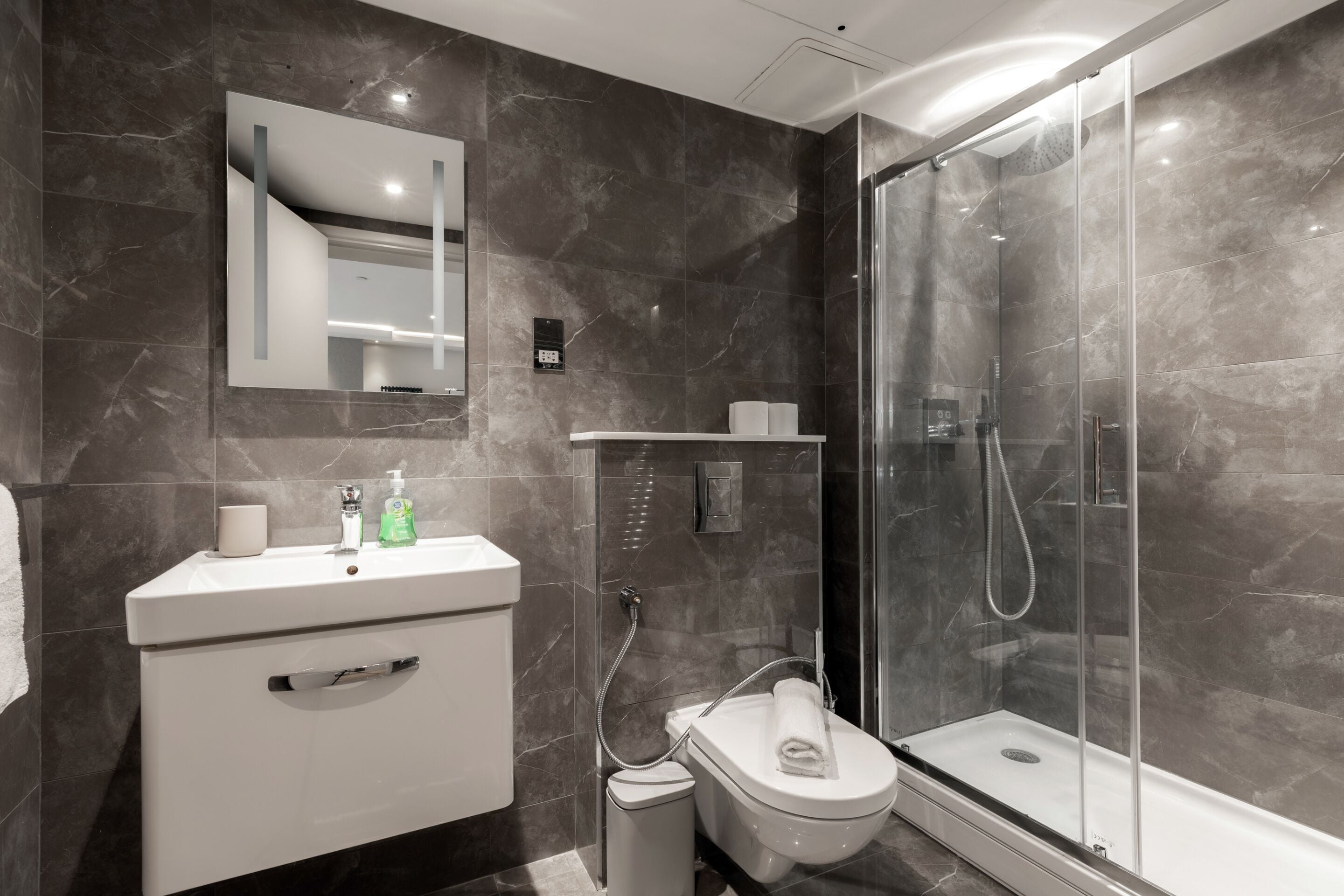 A modern bathroom with a marble wall, a white sink, a toilet, and a glass-enclosed shower.