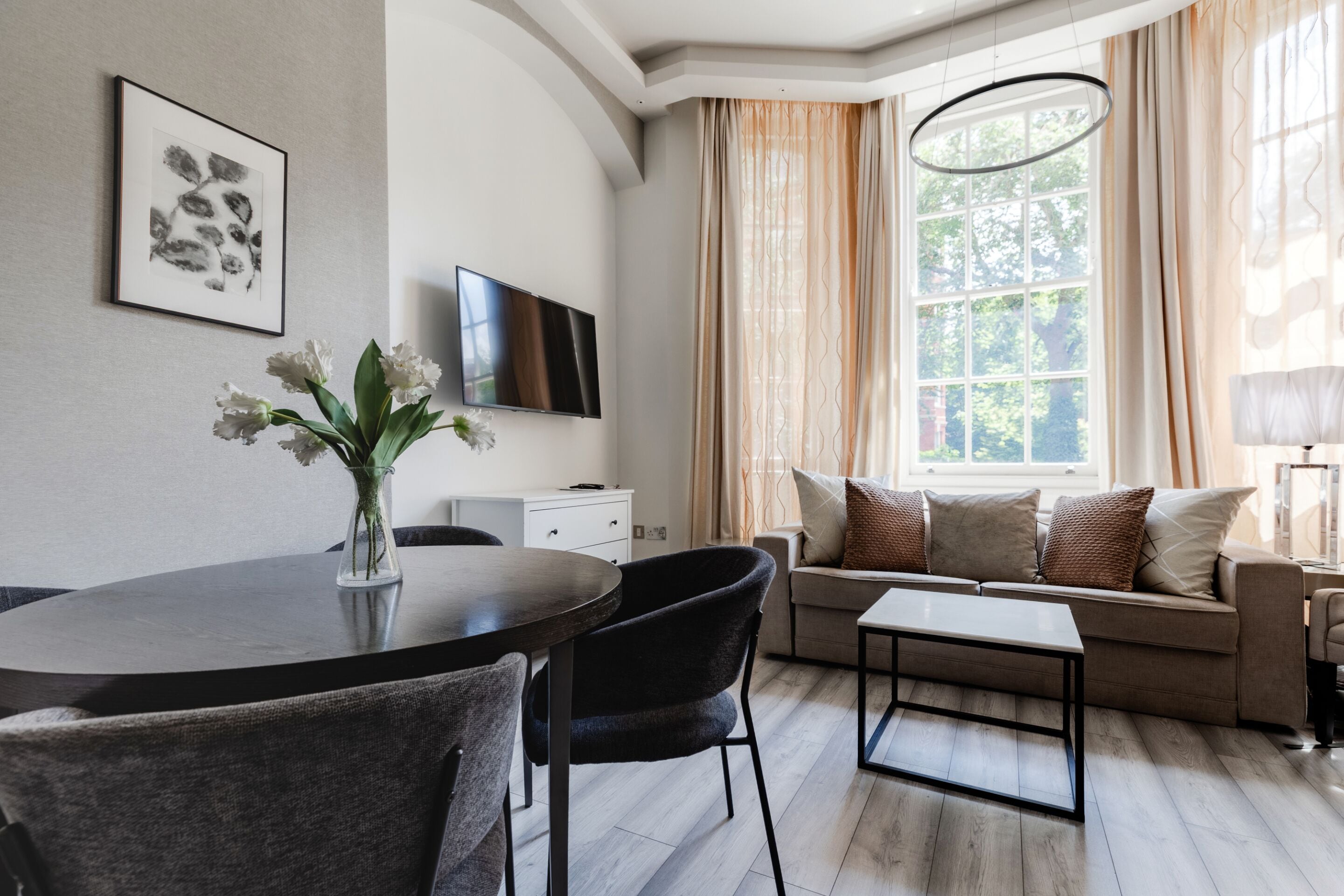A modern living room with a round dining table, black chairs, a sofa with cushions, a flat-screen TV, and a large window with curtains.