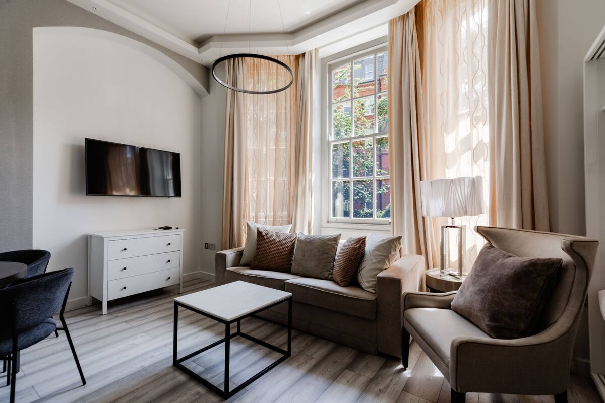 Un salon moderne avec un canapé, un fauteuil et une table basse. Un téléviseur à écran plat est fixé au mur, et une fenêtre avec des rideaux est visible.