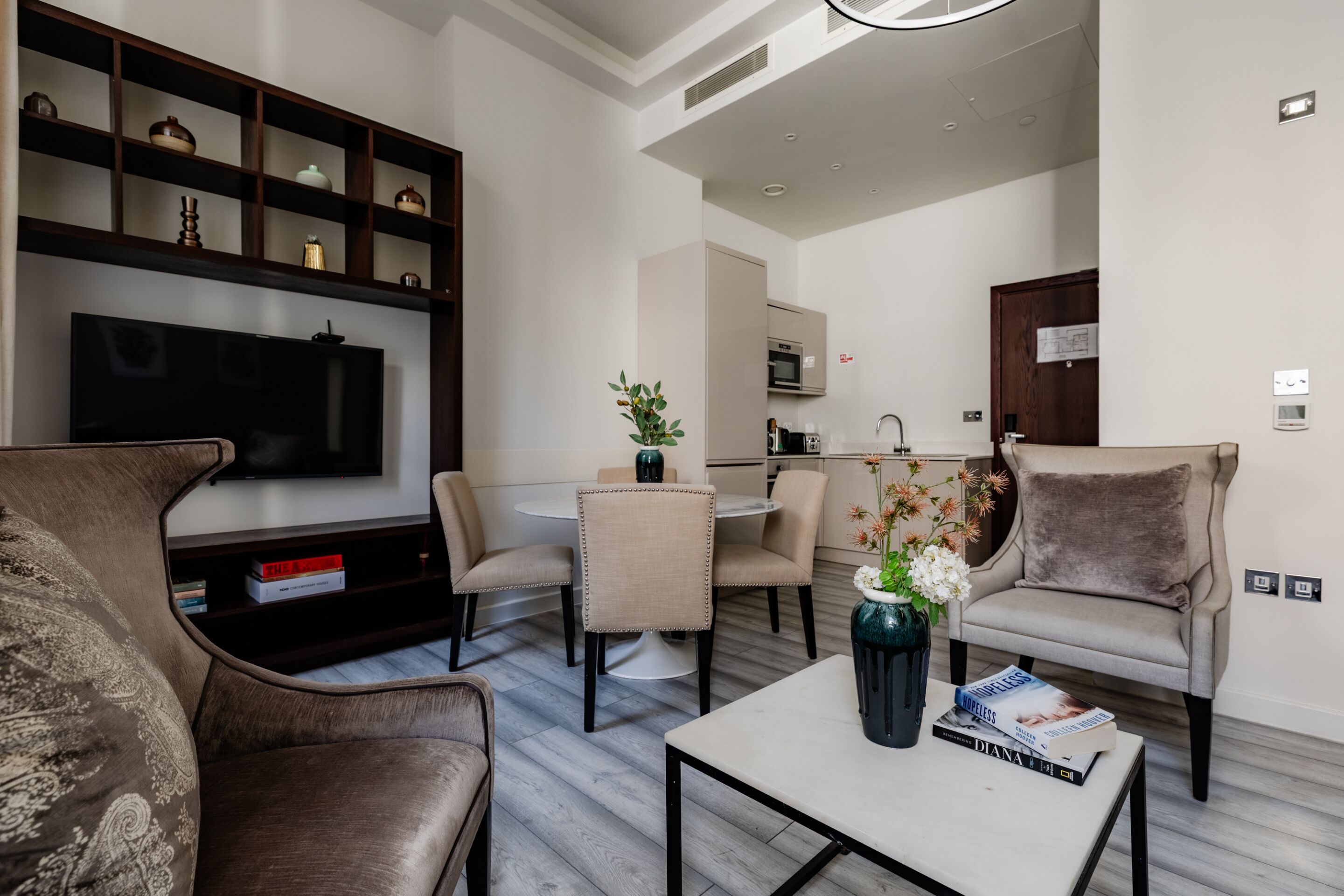 A modern living room with a flat-screen TV, comfortable seating, a dining area, and a kitchen in the background.