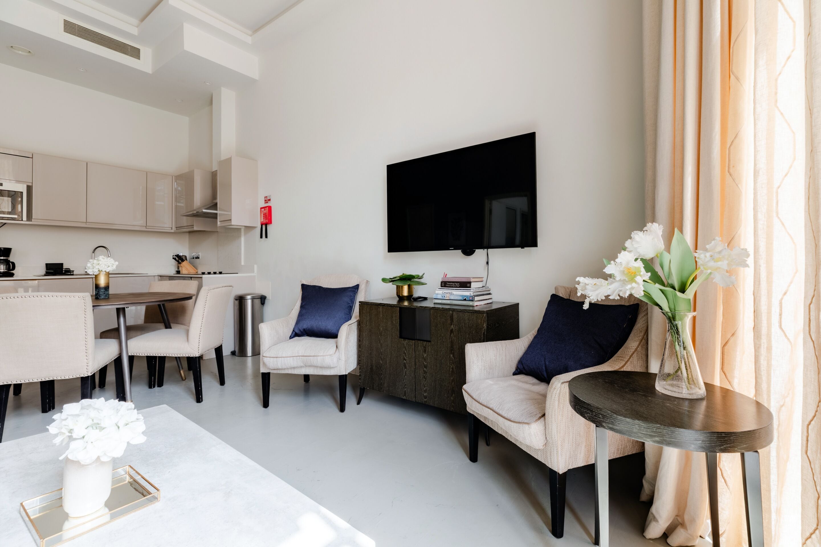 A modern living room with a dining area, a flat-screen TV mounted on the wall, and a vase with flowers on a side table.